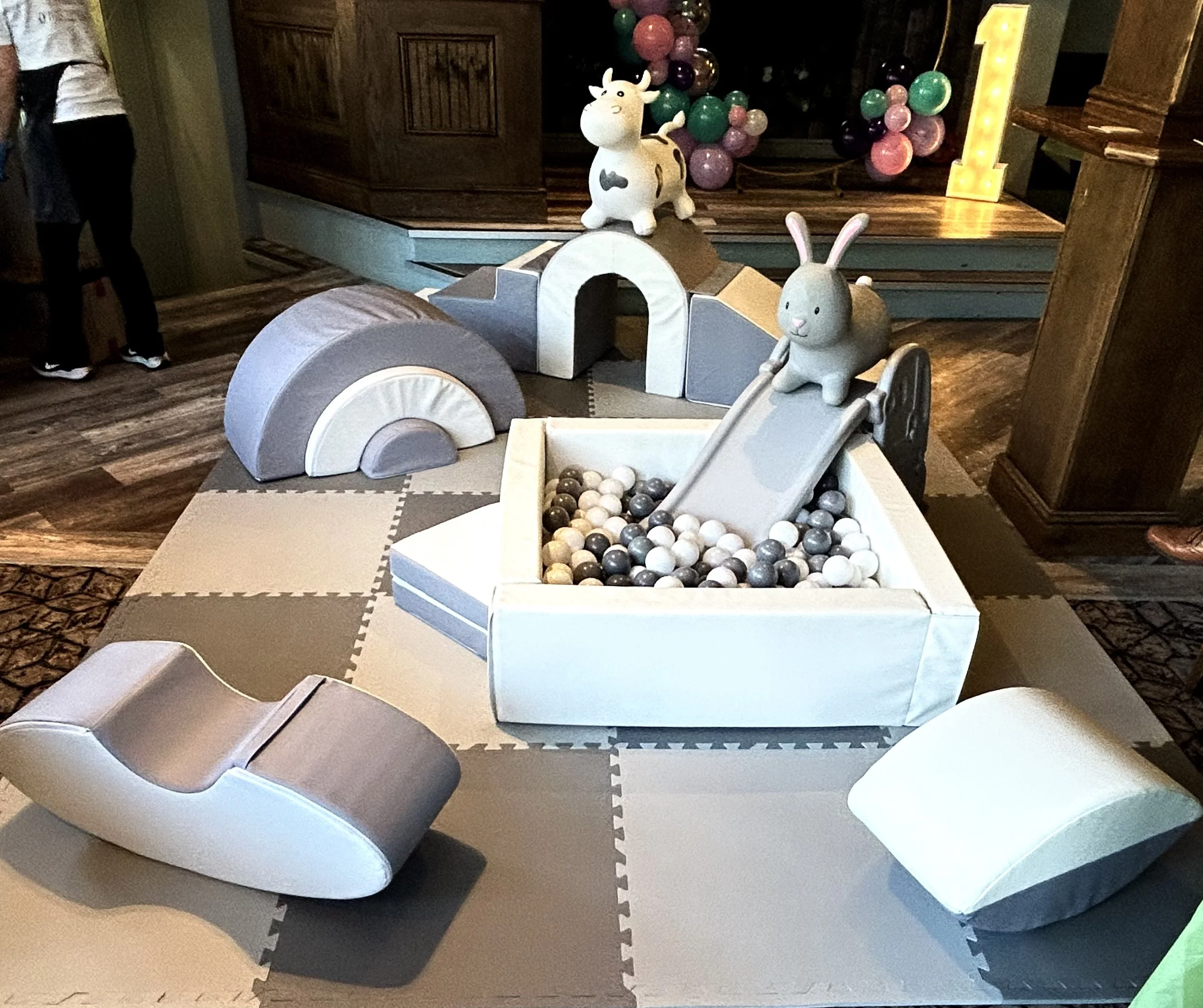 Children's indoor play area with soft foam equipment, a ball pit with black and white balls, and decorative farm animal toys, including a cow and a rabbit, on a foam mat floor.