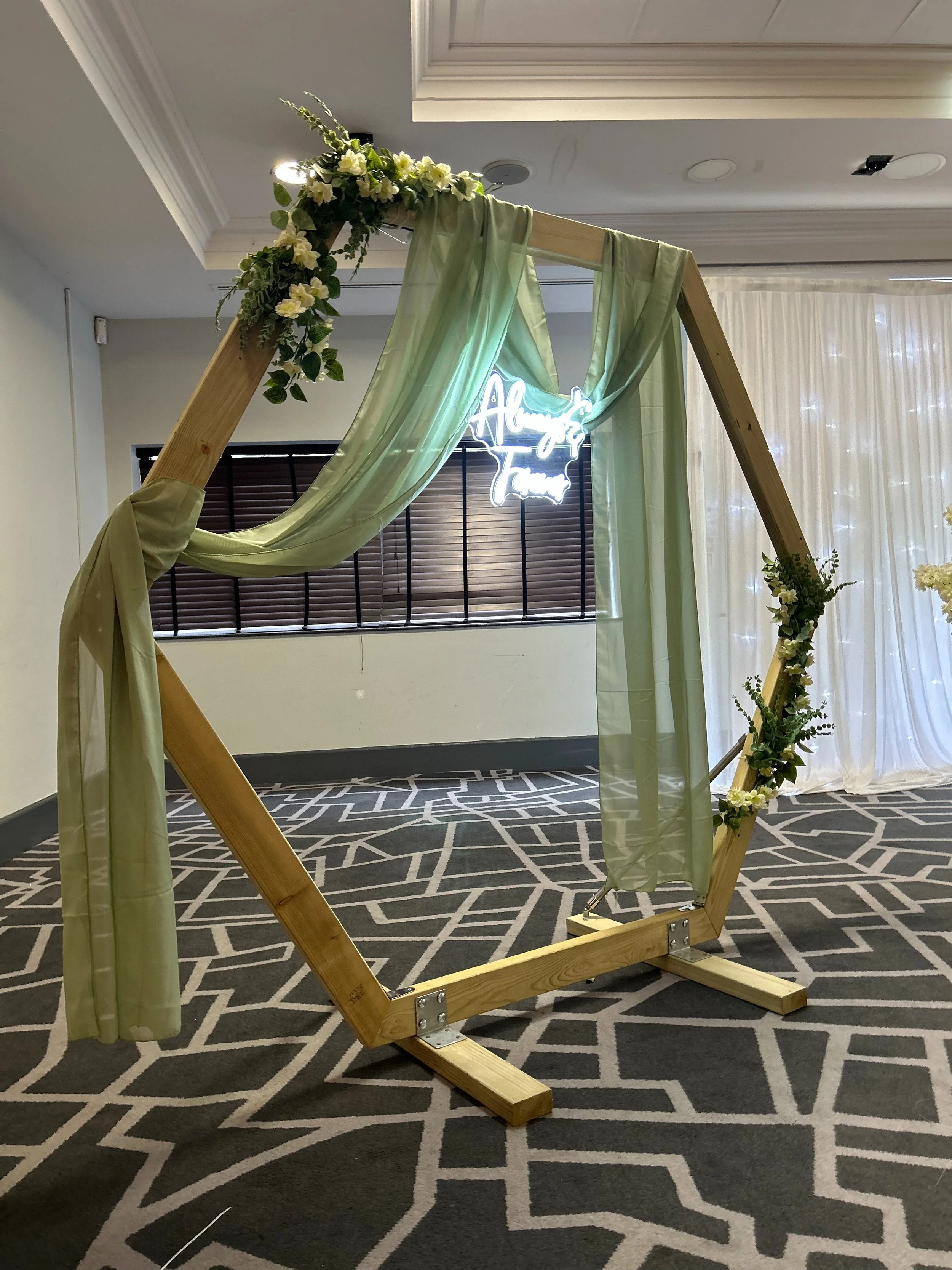 Wooden hexagonal arch decorated with green sheer fabric and white flowers, with a neon sign in the background that says 'Always & Forever'.