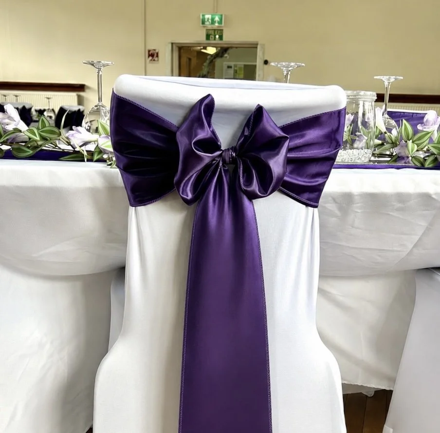 A white banquet chair decorated with a large purple satin bow at the back, part of a decorated table setup with glassware and floral arrangements, in a banquet hall