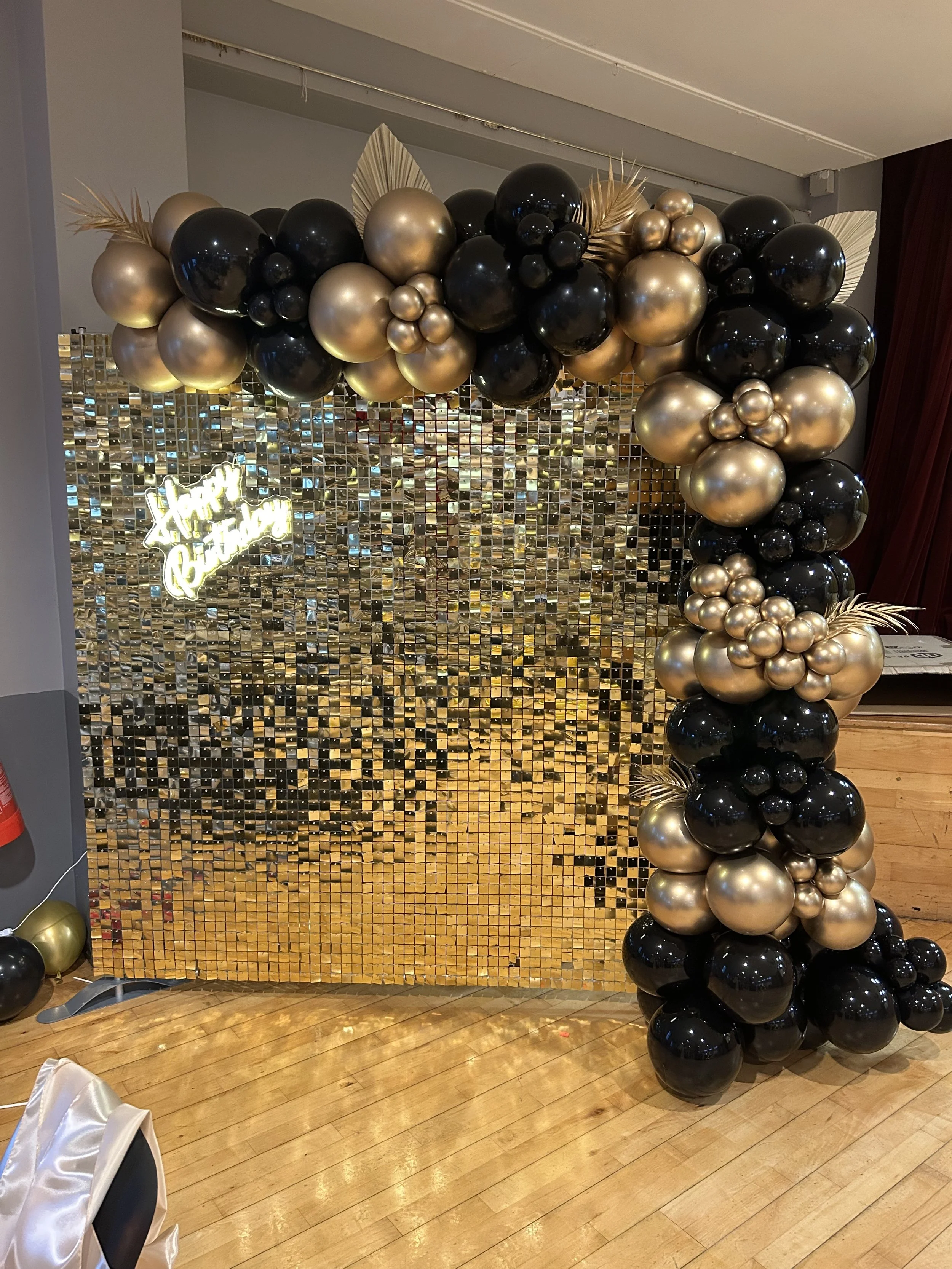 Decorative balloon arch with black and gold balloons, a gold mirrored backdrop, and a neon sign that says 'Happy Birthday' in cursive.