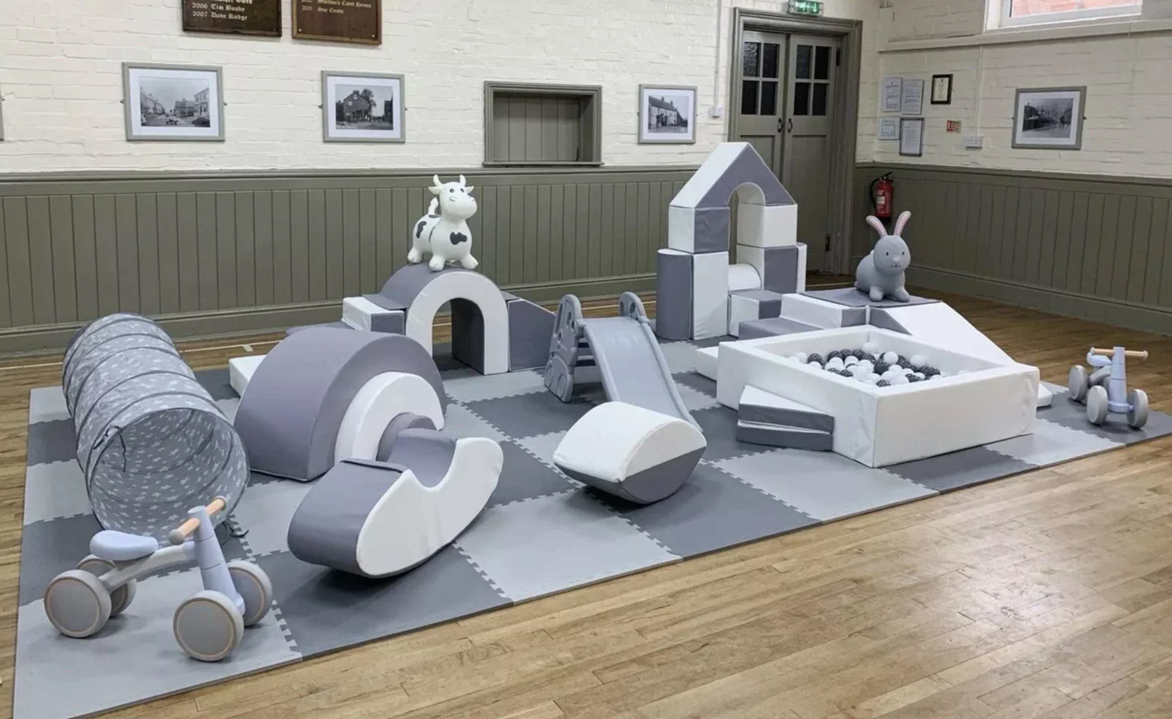 Gray and white children's play area with various toys and structures, including a tunnel, a slide, animal-shaped toys, a ball pit, and a tricycle, set up on a soft mat in an indoor space with framed photos and plaques on the wall.