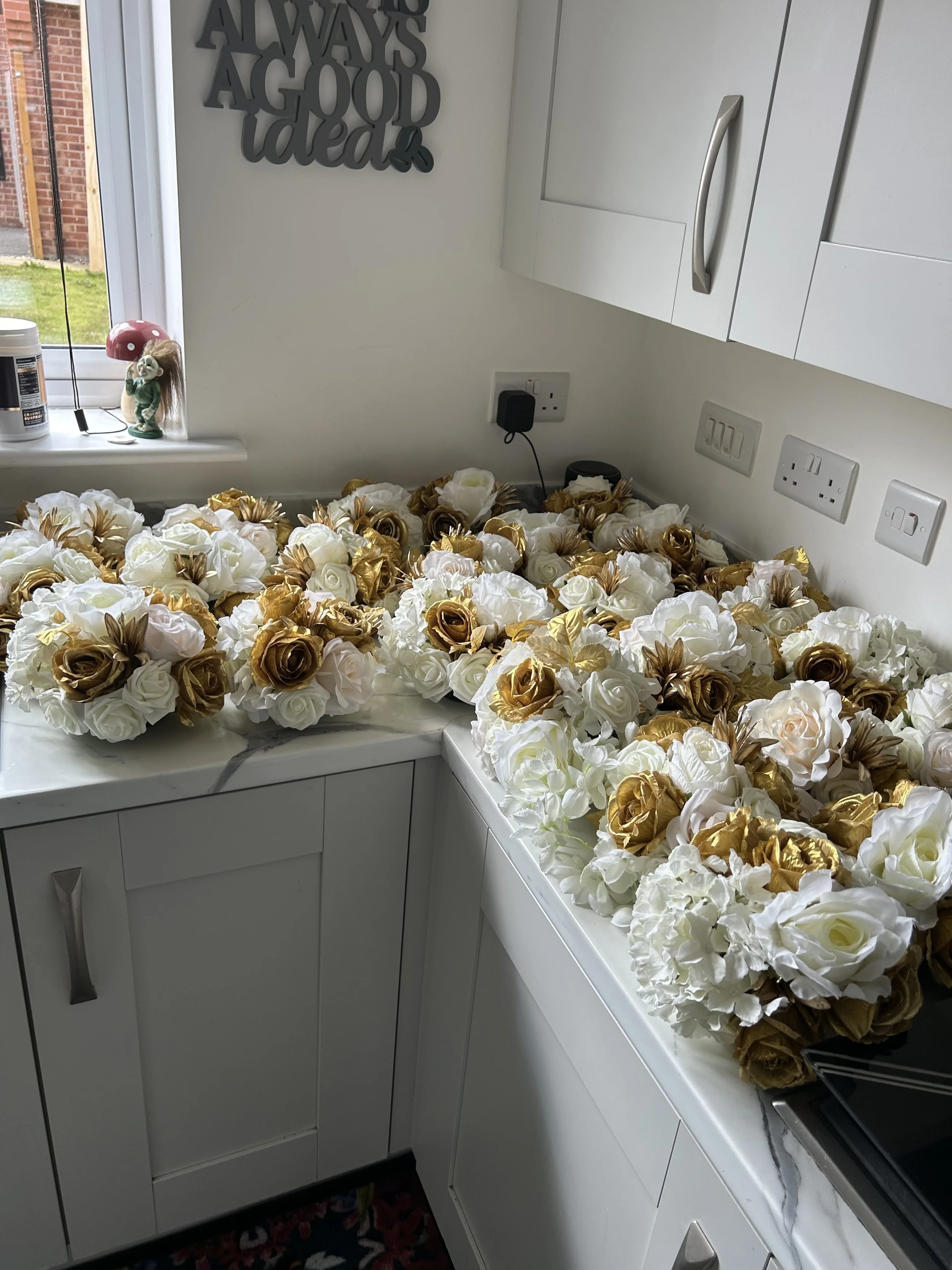 A kitchen countertop covered with numerous white, gold, and bronze artificial flower bouquets. A small window with a red mushroom-shaped lamp and a decorative doll are visible in the background. There is a wall sign that reads "Always a good idea" an