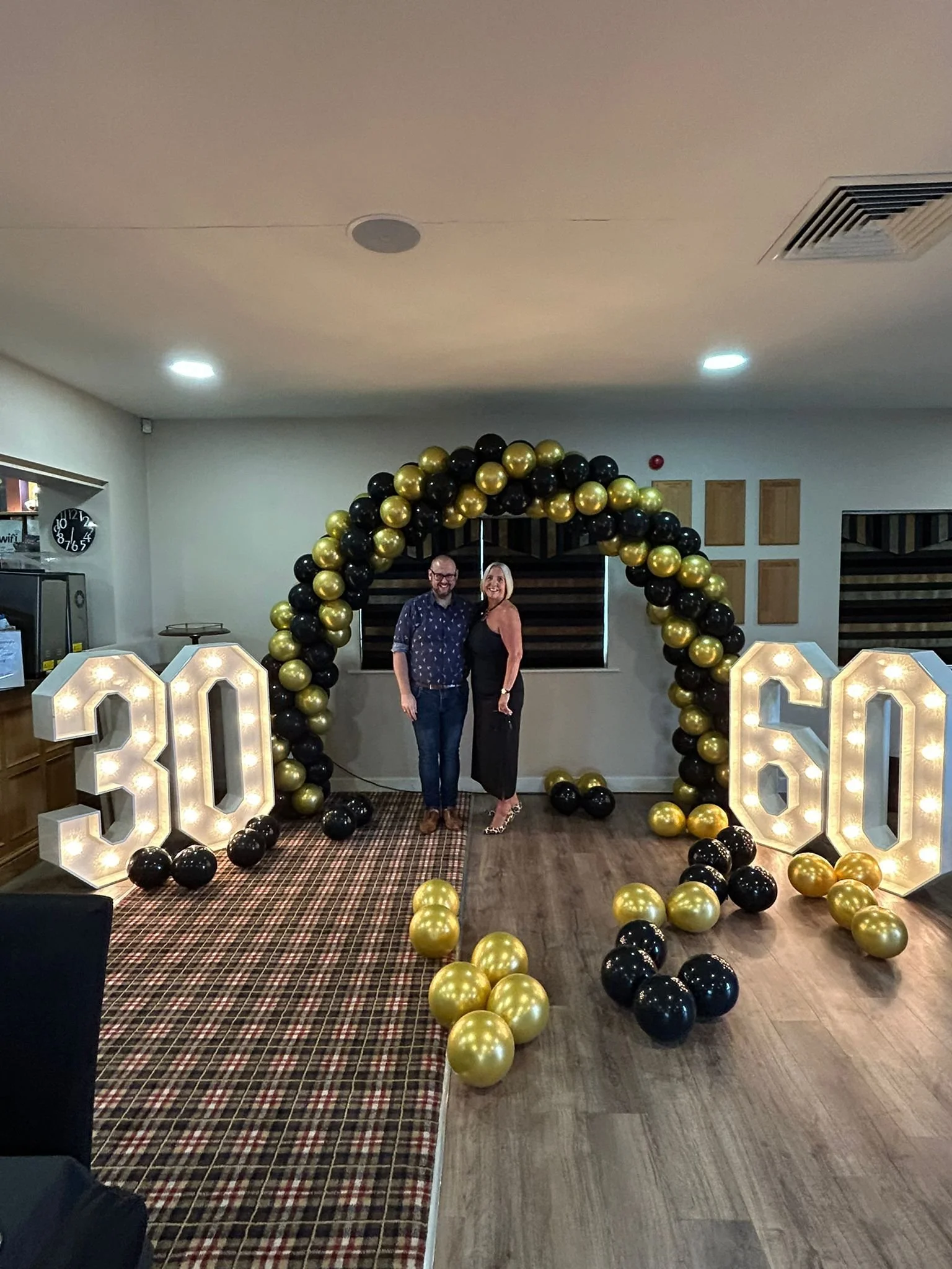 A celebrating 30th and 60th birthday party with two people standing in front of a balloon arch and illuminated numbers 30 and 60.