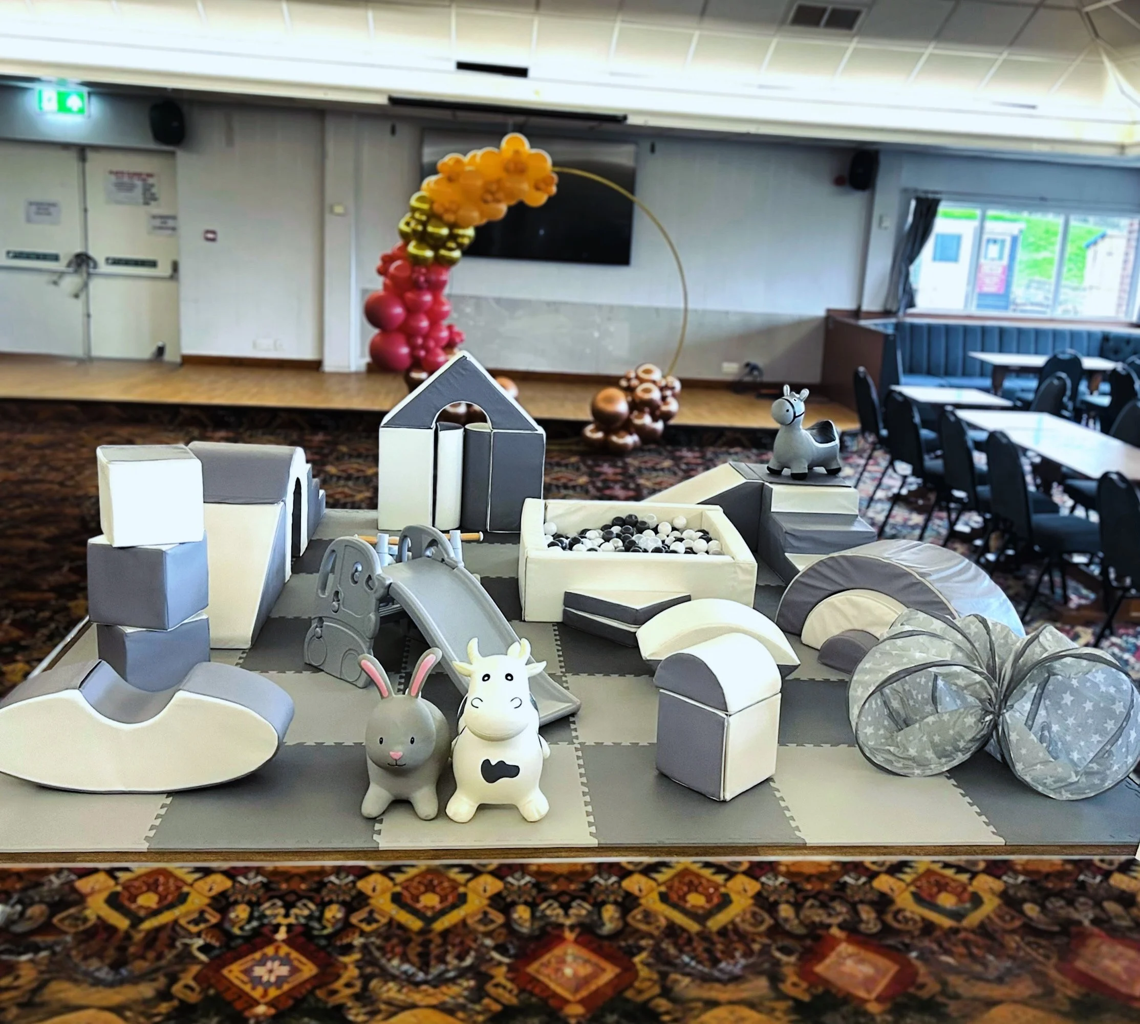 Children's soft play area with rabbit and unicorn toys, ball pit, slide, and various cushioned structures, set up in a banquet hall with tables and balloon decoration in the background.