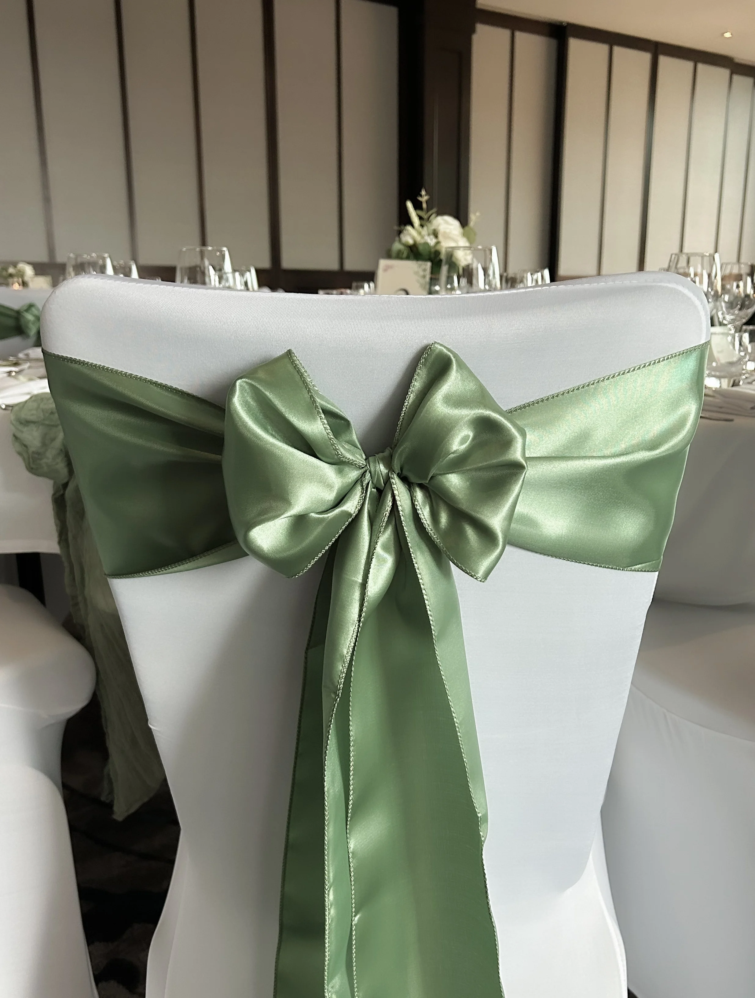 A white banquet chair decorated with a large, shiny light green satin bow tied at the back. The table behind the chair is set with wine glasses and floral centerpieces.