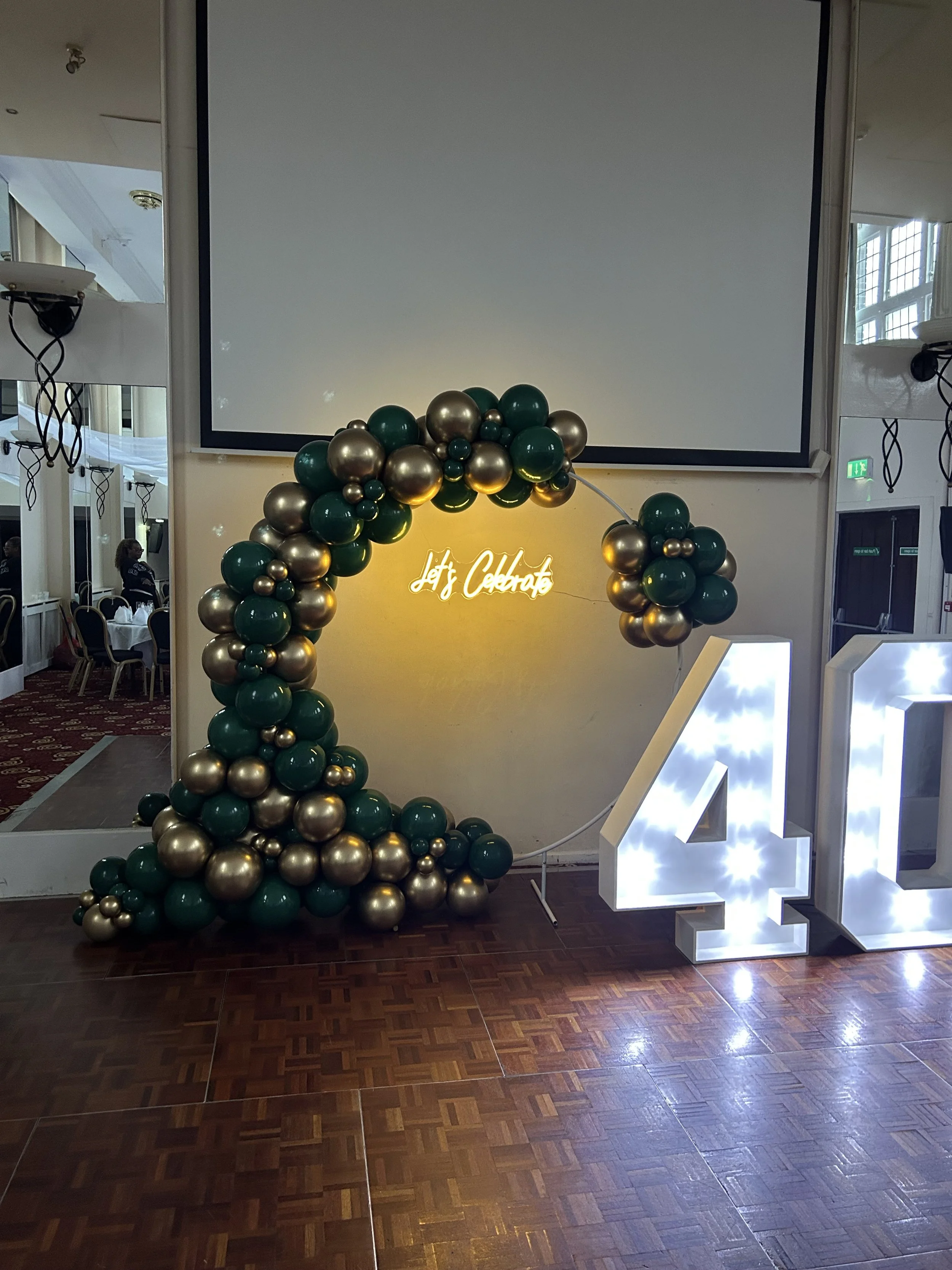 Party decoration with a large balloon arrangement in green and gold, a neon sign saying "Let's Celebrate," and illuminated letters "4" and "0" for a 40th birthday celebration.