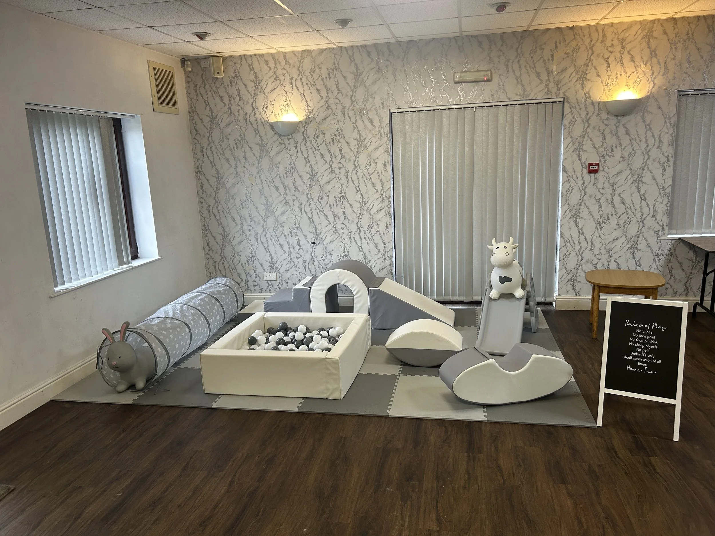 Indoor play area with soft toys, ball pit, and slides for children, located in a room with patterned wallpaper, wooden floor, and window with blinds.