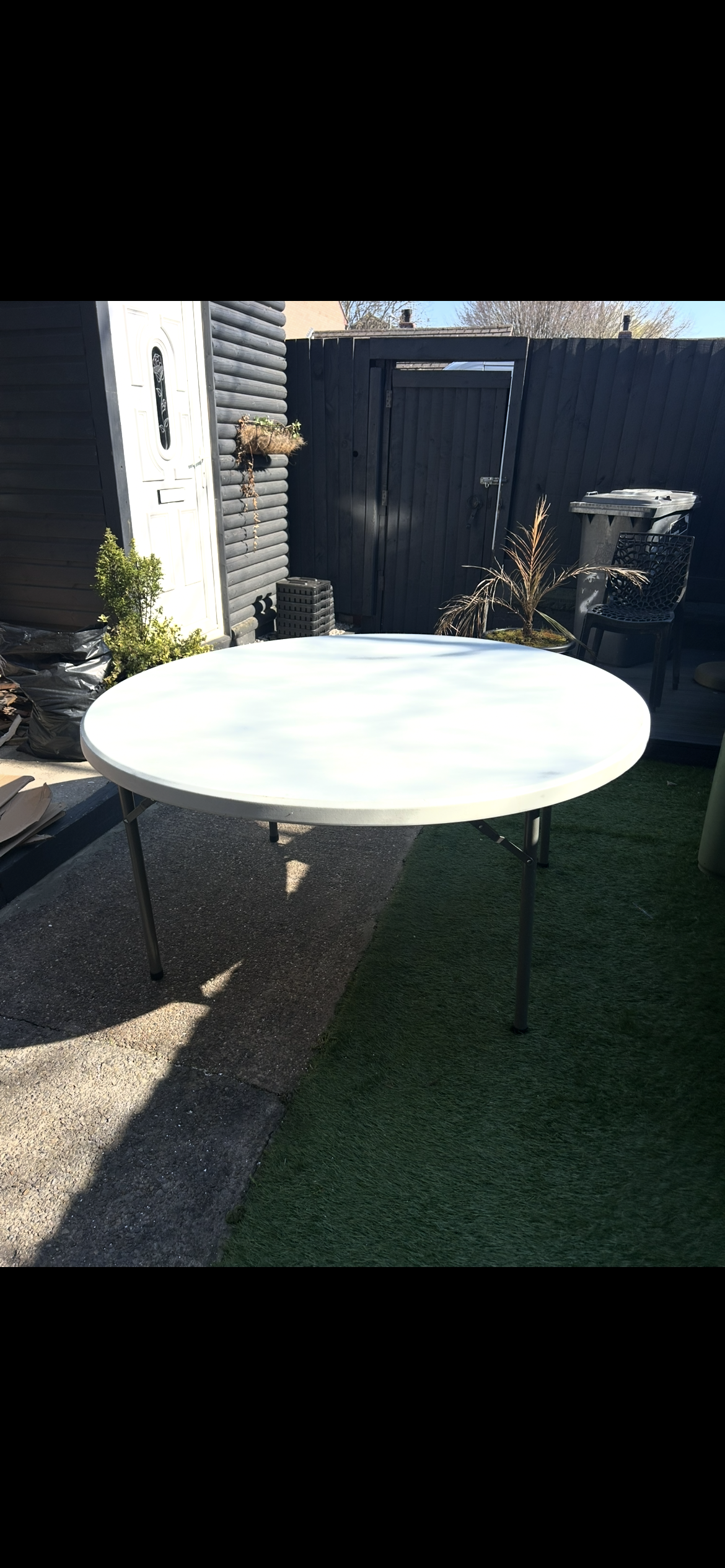 Outdoor backyard scene with a round white plastic table, black fence, garden decorations, a black chair with lattice back, recycling bin, small plants, and a patch of artificial turf.