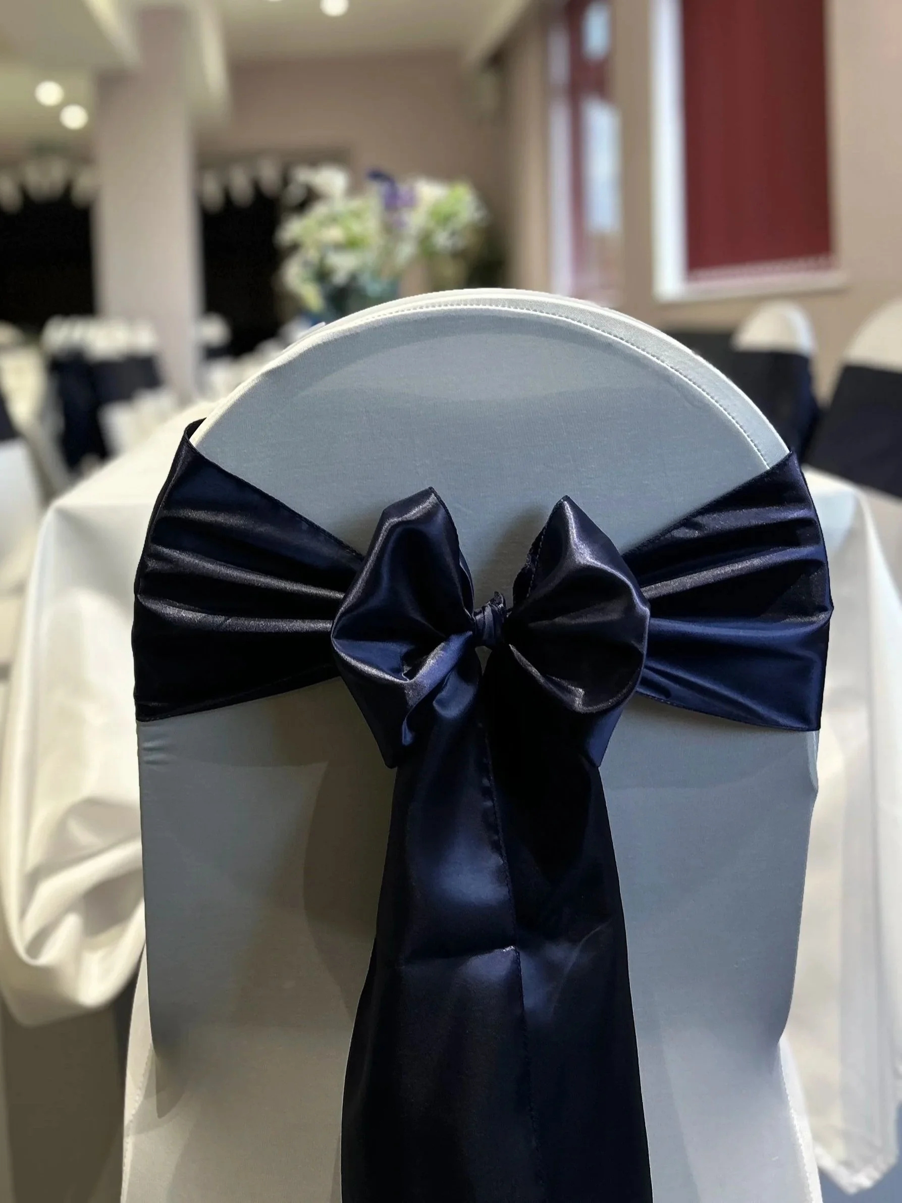 A white chair with a navy blue satin bow tied around the backrest, decorated as a wedding or event centerpiece. In the background, there are other chairs, a table with a white tablecloth, and a vase with flowers.