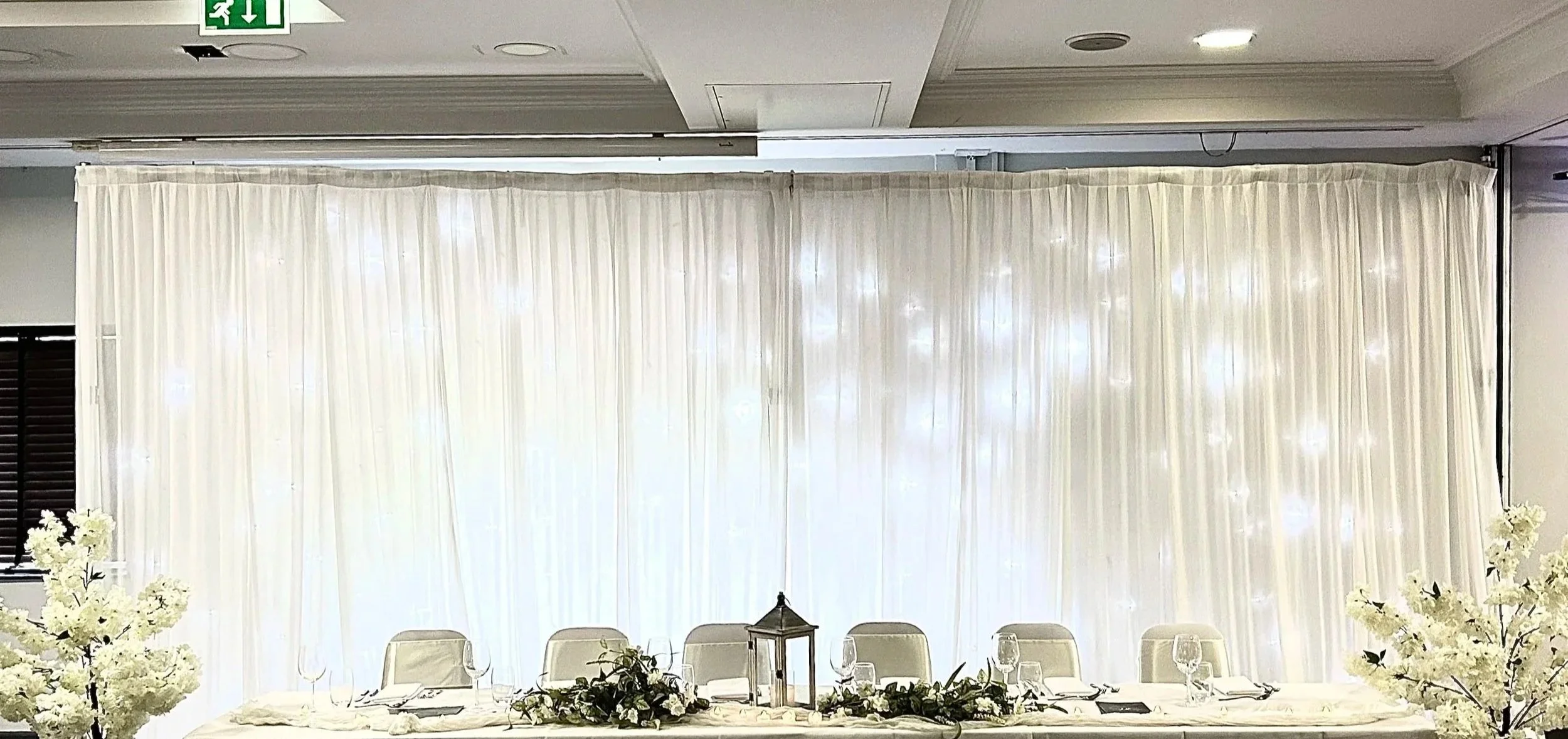 Elegant decorated banquet table with white chairs, a lantern centerpiece, greenery, and white flower arrangements in front of a sheer white curtain backdrop.