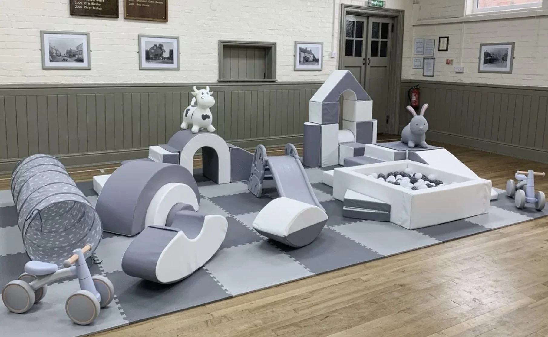 Indoor children's play area with soft, padded gray and white structures, including a tunnel, slide, ball pit, and animal-shaped toys such as a cow and a rabbit. The area has wood flooring and framed photos on the wall.
