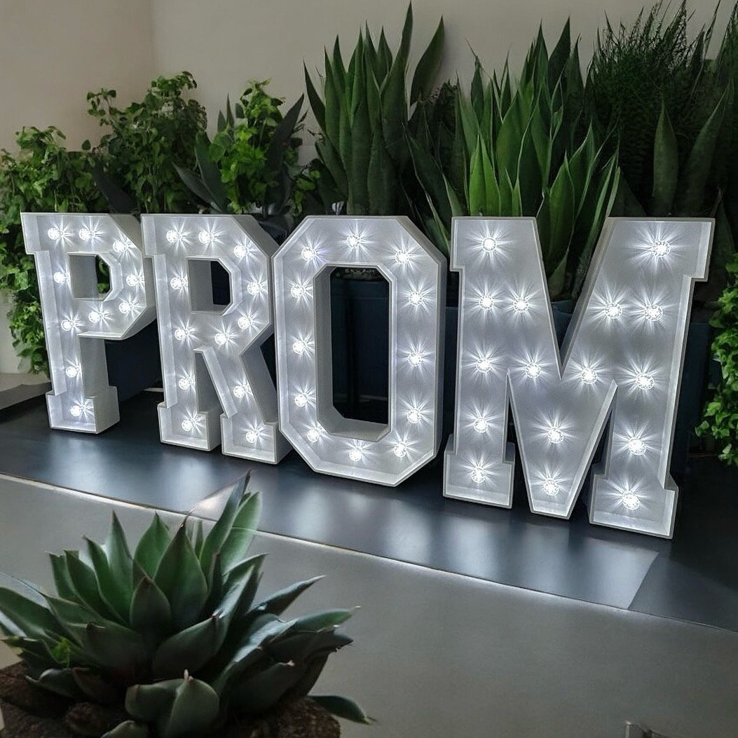 Decorative marquee sign spelling 'PROM' with bright white lights, placed in front of green potted plants on a black surface.