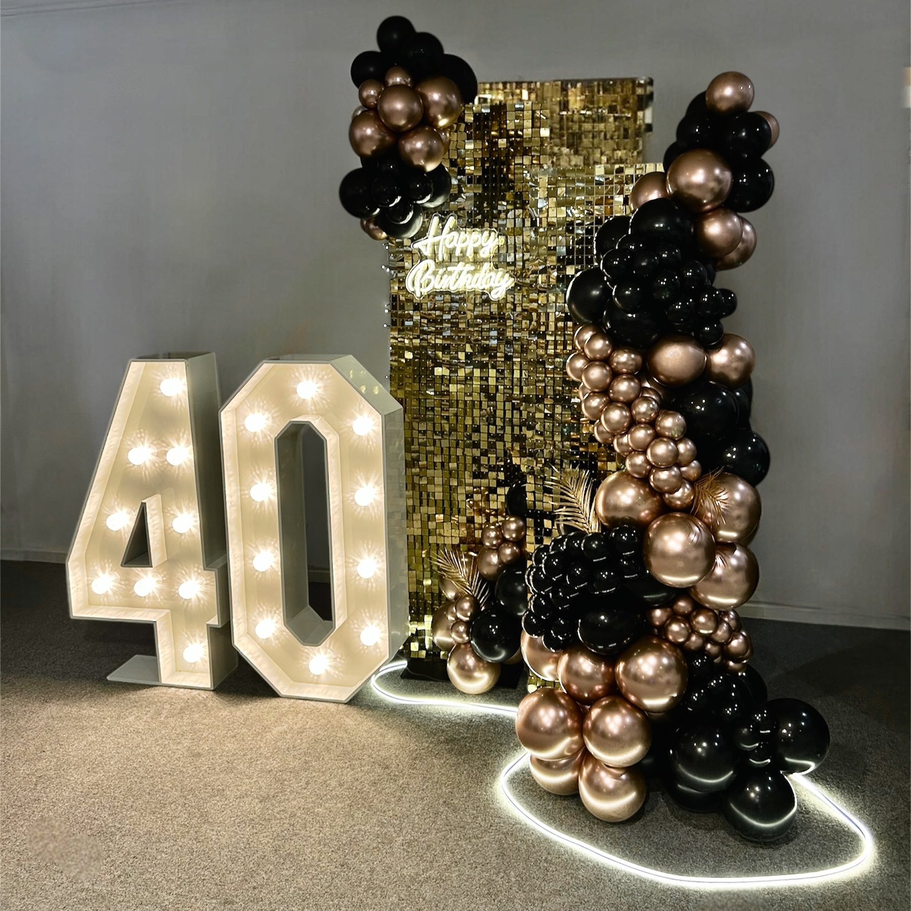Decorative birthday display with large illuminated number 40, black and gold balloons, a gold sequin backdrop, a neon sign that says 'Happy Birthday,' and a circular LED light on the floor outlining the display.