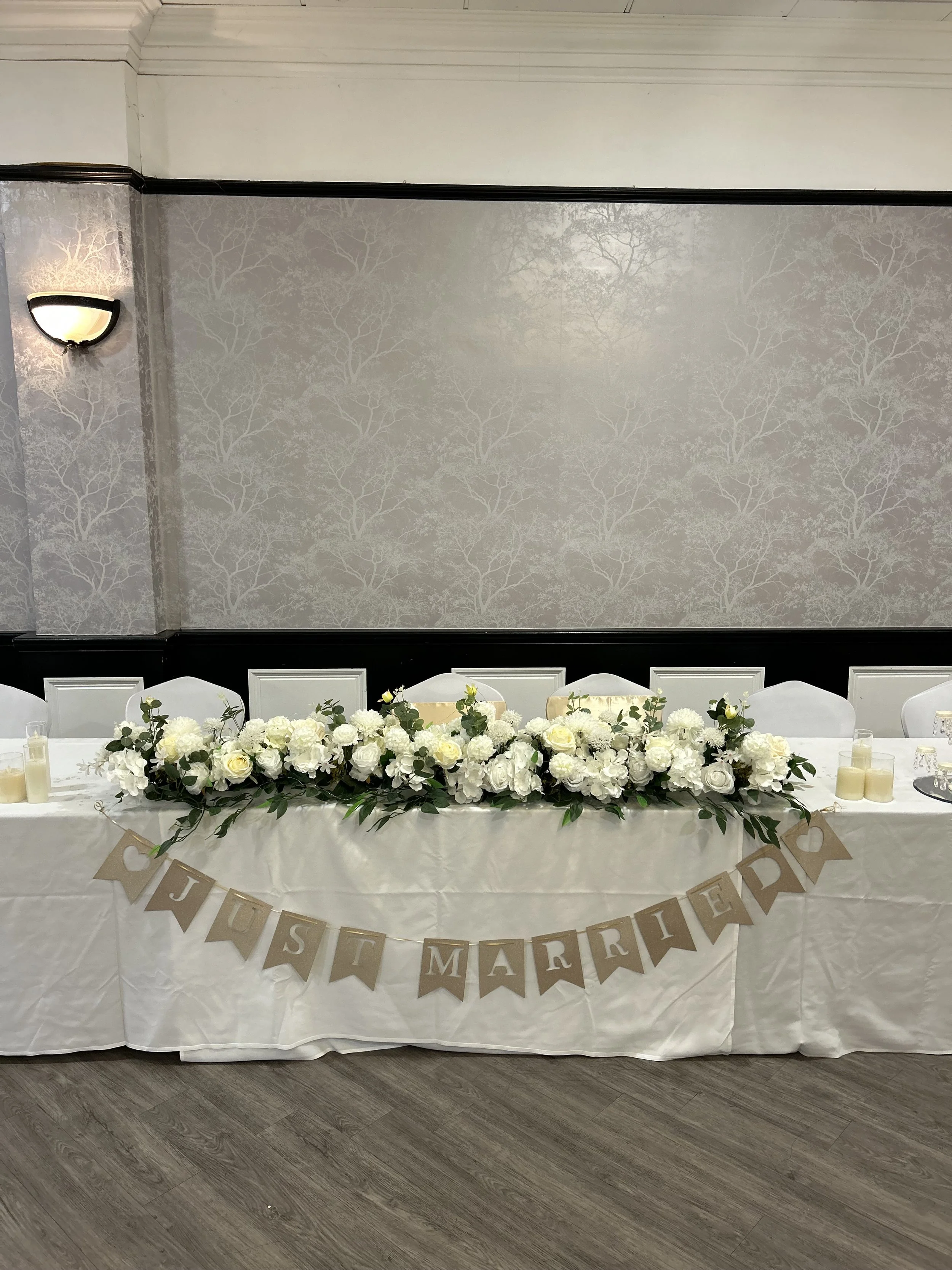 A long rectangular table decorated with a cascading floral arrangement of white roses, white hydrangeas, and greenery. The table has several white chairs, and a 'Just Married' banner with individual pennants hanging below. The backdrop features a gray patterned wall with tree motifs, and a wall sconce light fixture.