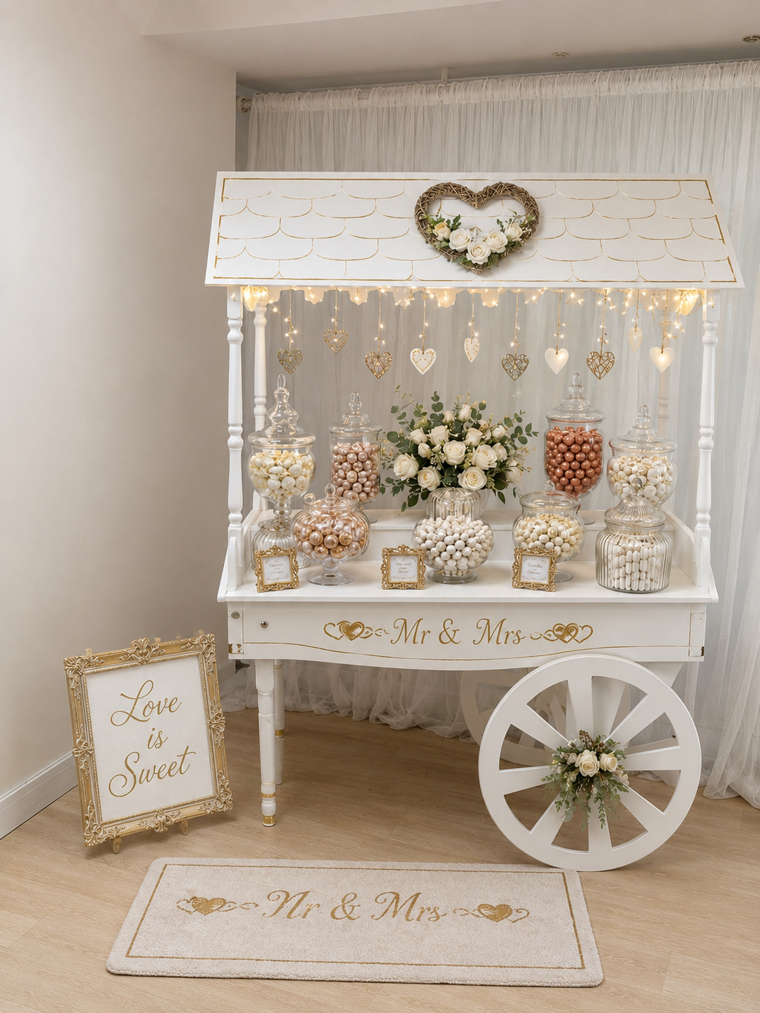 Decorative candy cart with jars of white and pink candies, white roses, and hanging heart ornaments, with a sign reading 'Love is Sweet' and 'Mr & Mrs'.