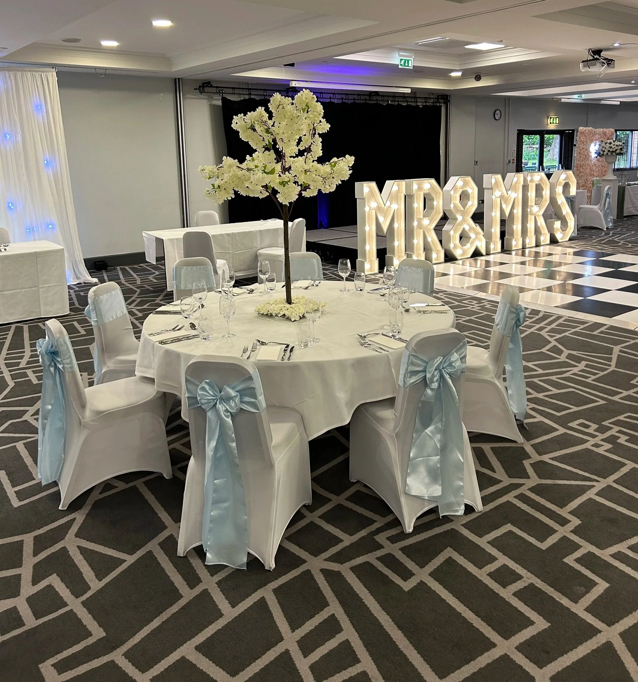 Event hall decorated with white drapes, tables, and chairs. A tall floral arrangement with white flowers is on a table. Large illuminated letters spell M&X&D&Y& S.