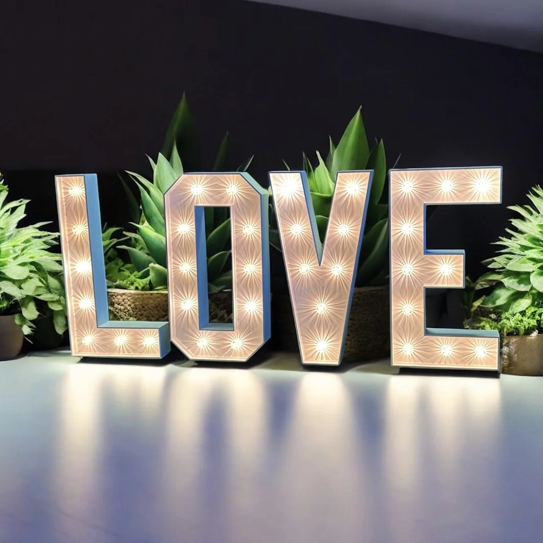 Large illuminated letters spelling 'LOVE' in a decorated room with patterned curtains, wall sconces, and a patterned carpet, above a radiator near an exit sign.