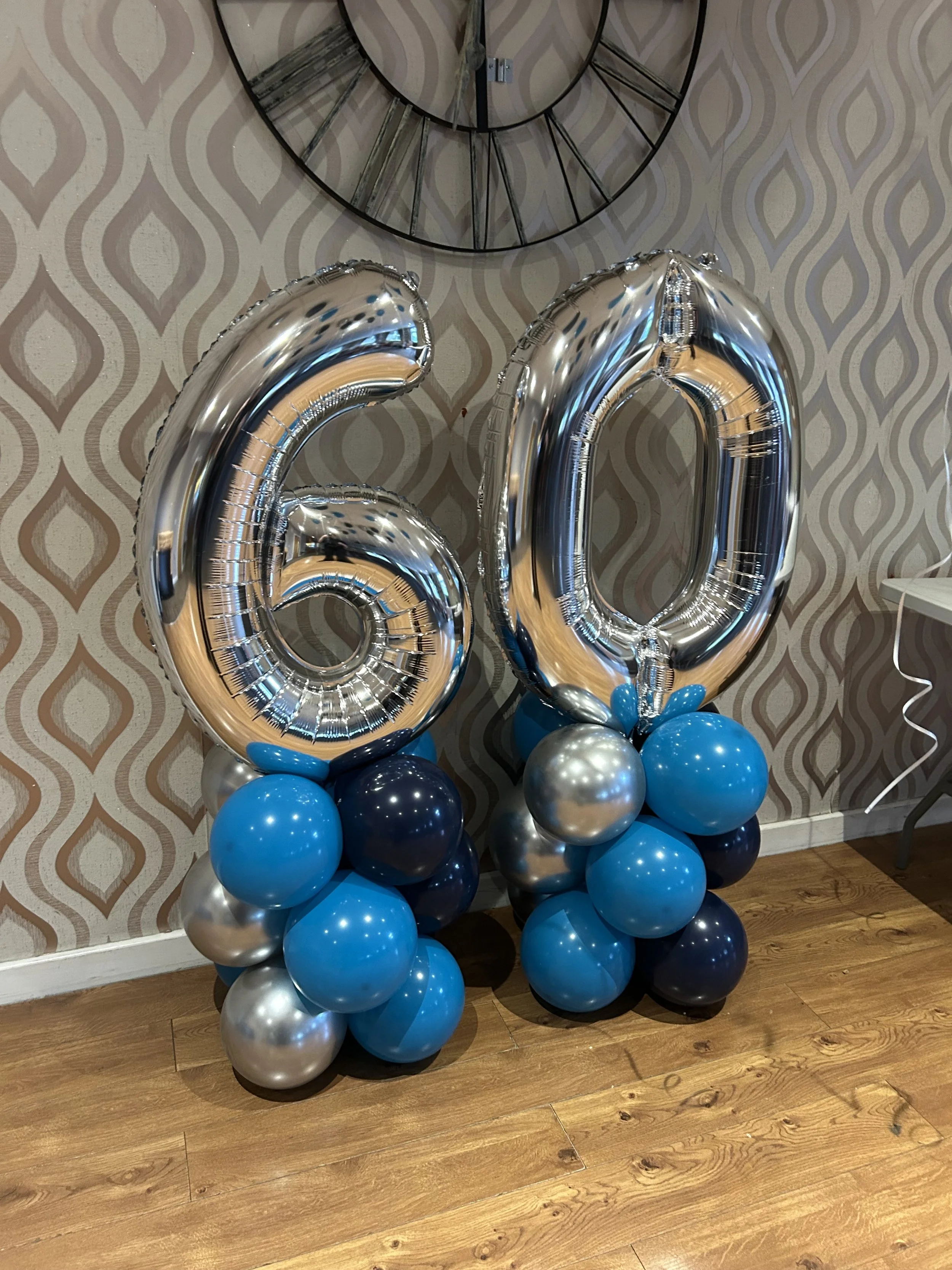 Silver and blue balloons forming the number 60, placed against a wallpaper with a diamond pattern, and a wall clock above them.
