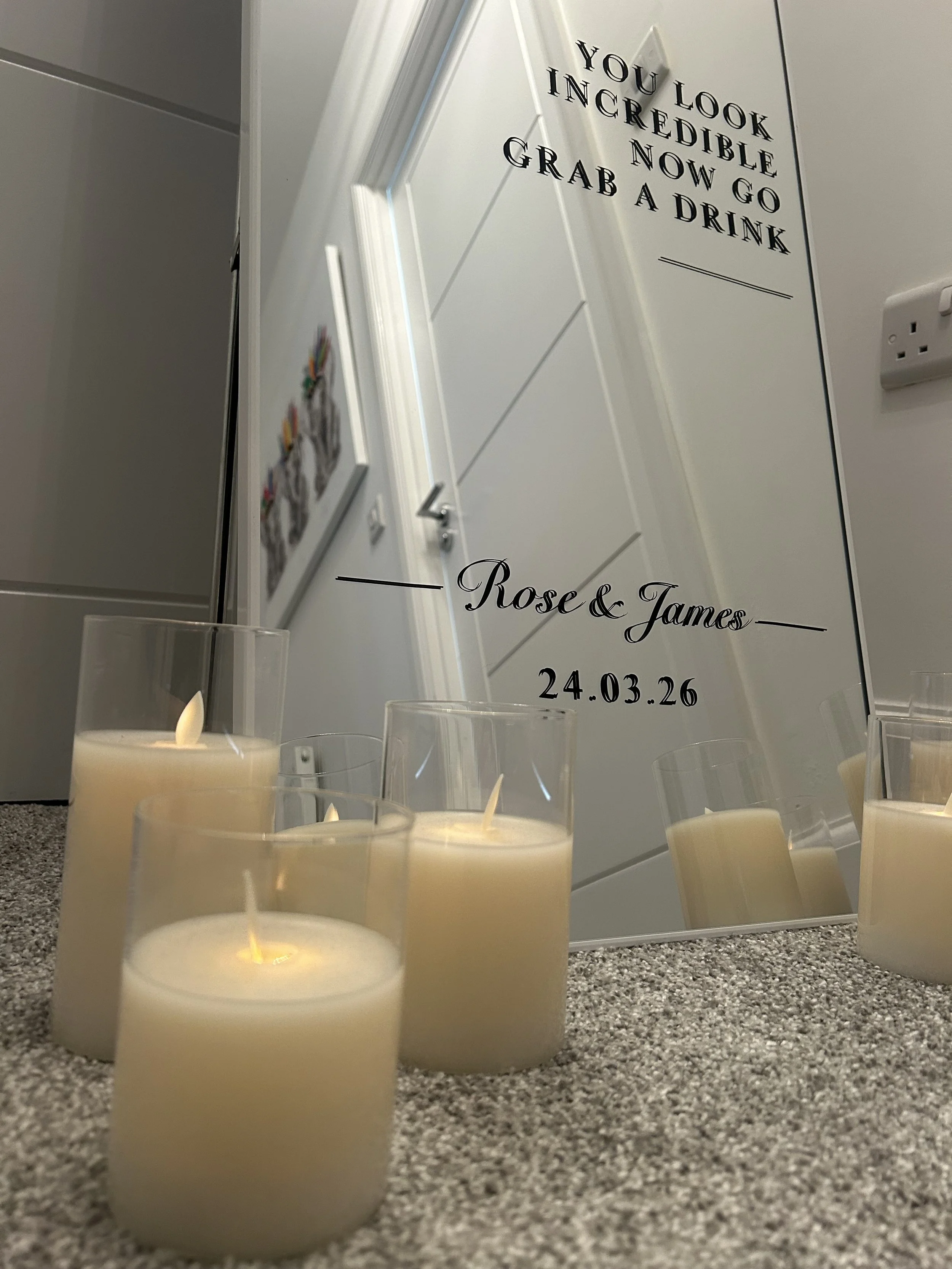 A mirror with decorative text and a date, surrounded by four lit beige candles, placed on a carpeted floor in front of a white door.
