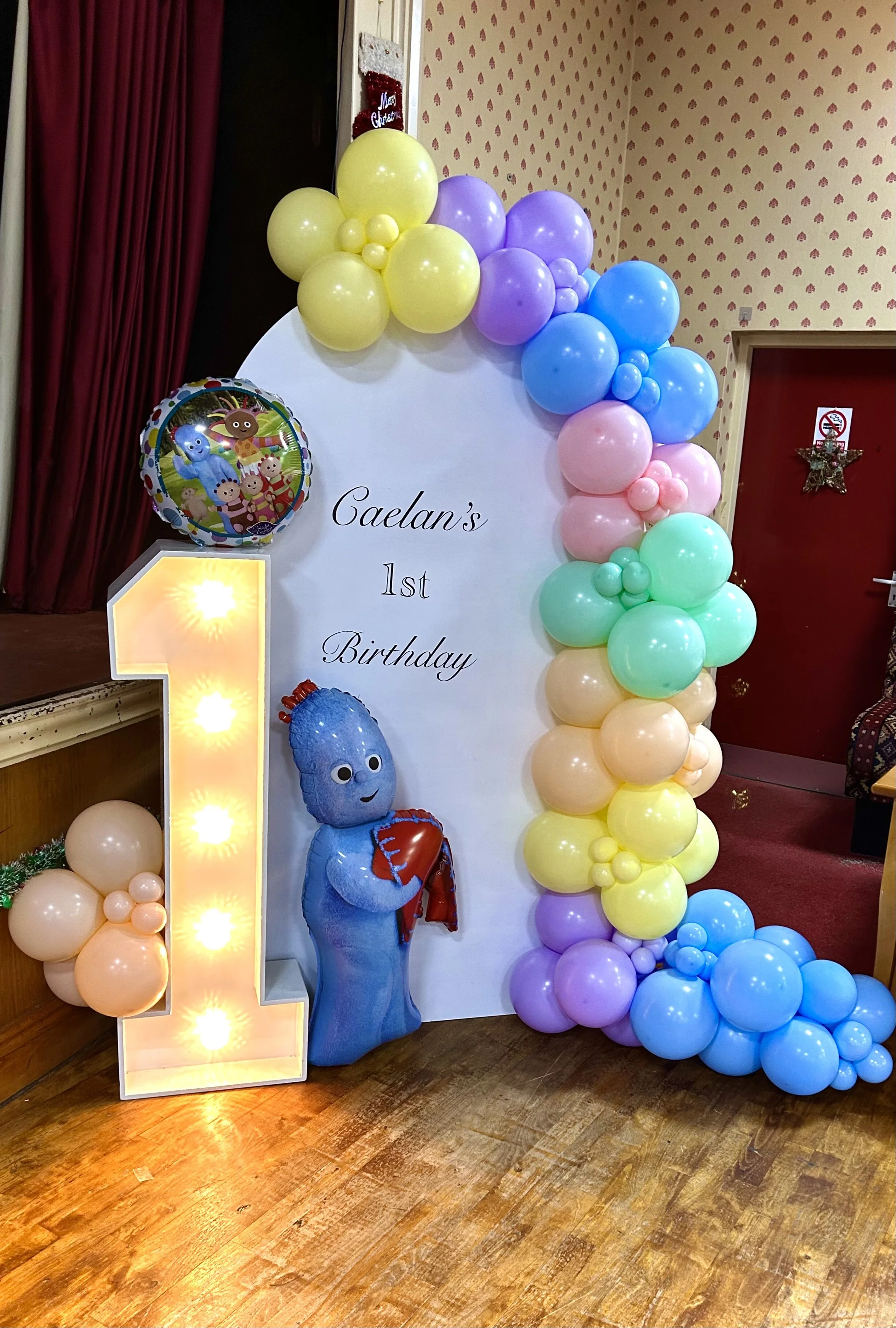 Decorative display for a child's first birthday, featuring a white board with text, colorful balloons arranged around the board, an illuminated number one, a character balloon from a children’s show, and a themed balloon with characters from the show
