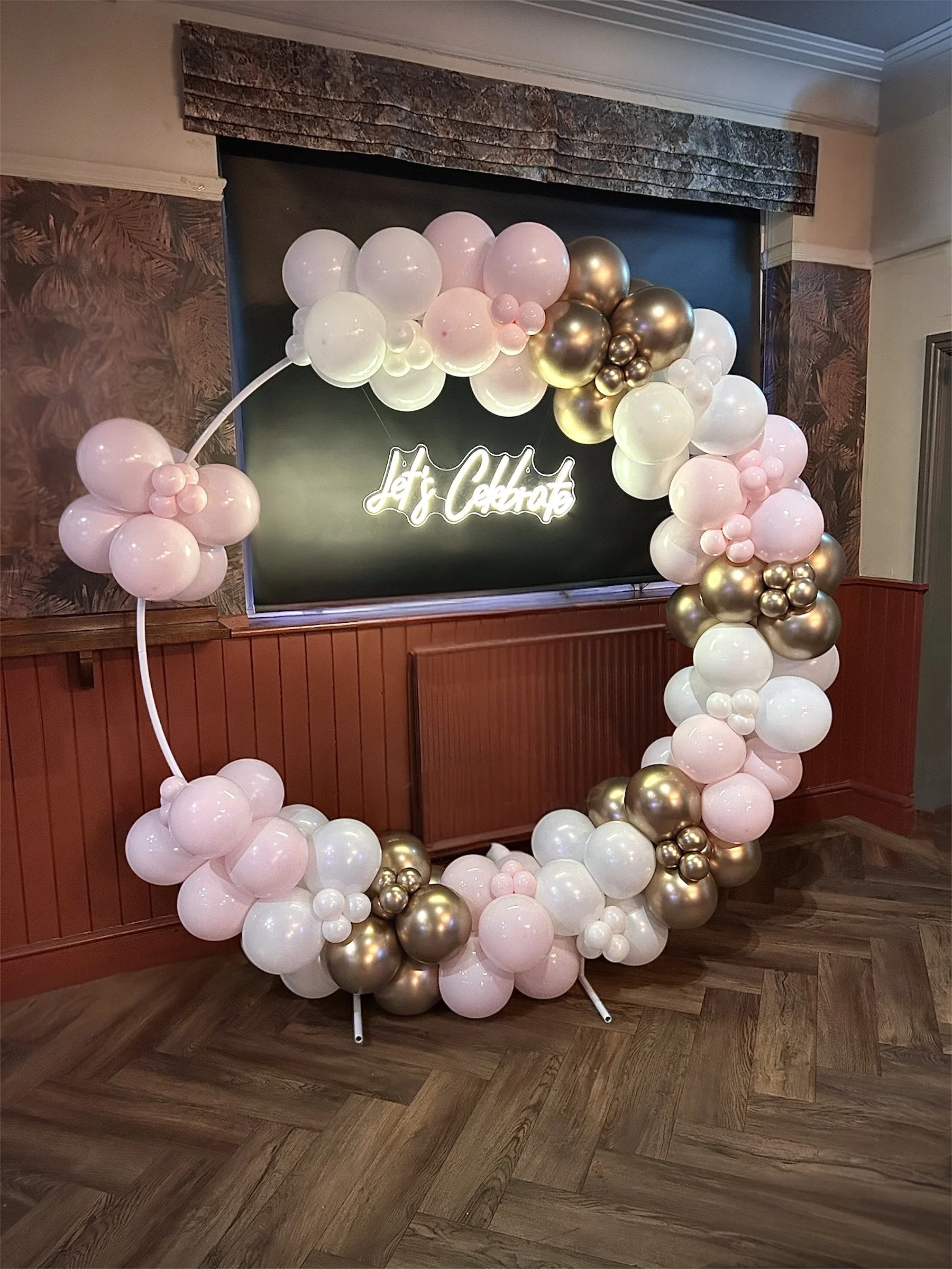 Decorative balloon arch with pink, white, and gold balloons, with a neon sign that says 'Let's Celebrate' in the background.