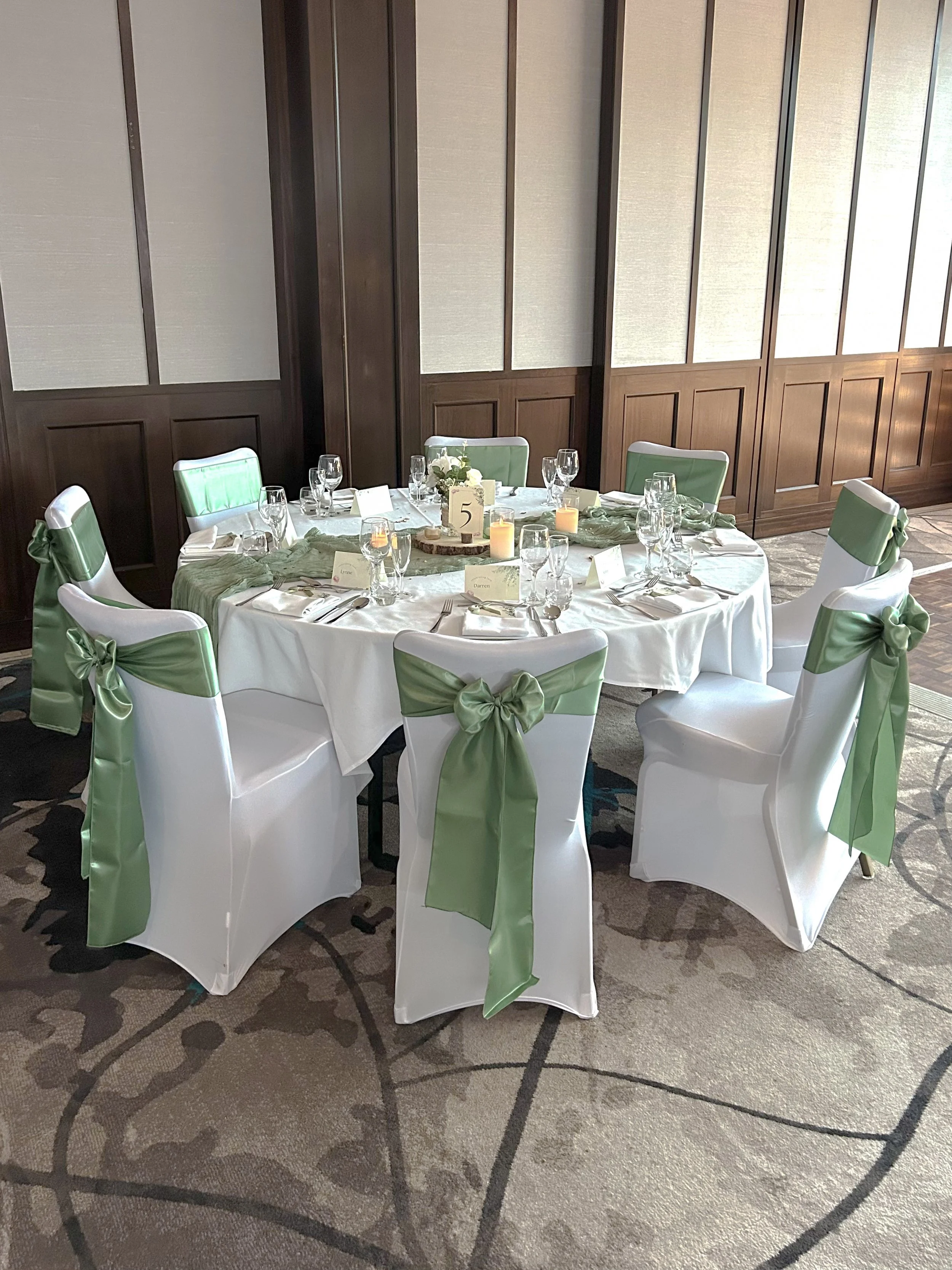 A round banquet table decorated for an event, with white tablecloth, green chair sashes, candles, and place settings, situated on a patterned carpeted floor with wood-paneled walls.