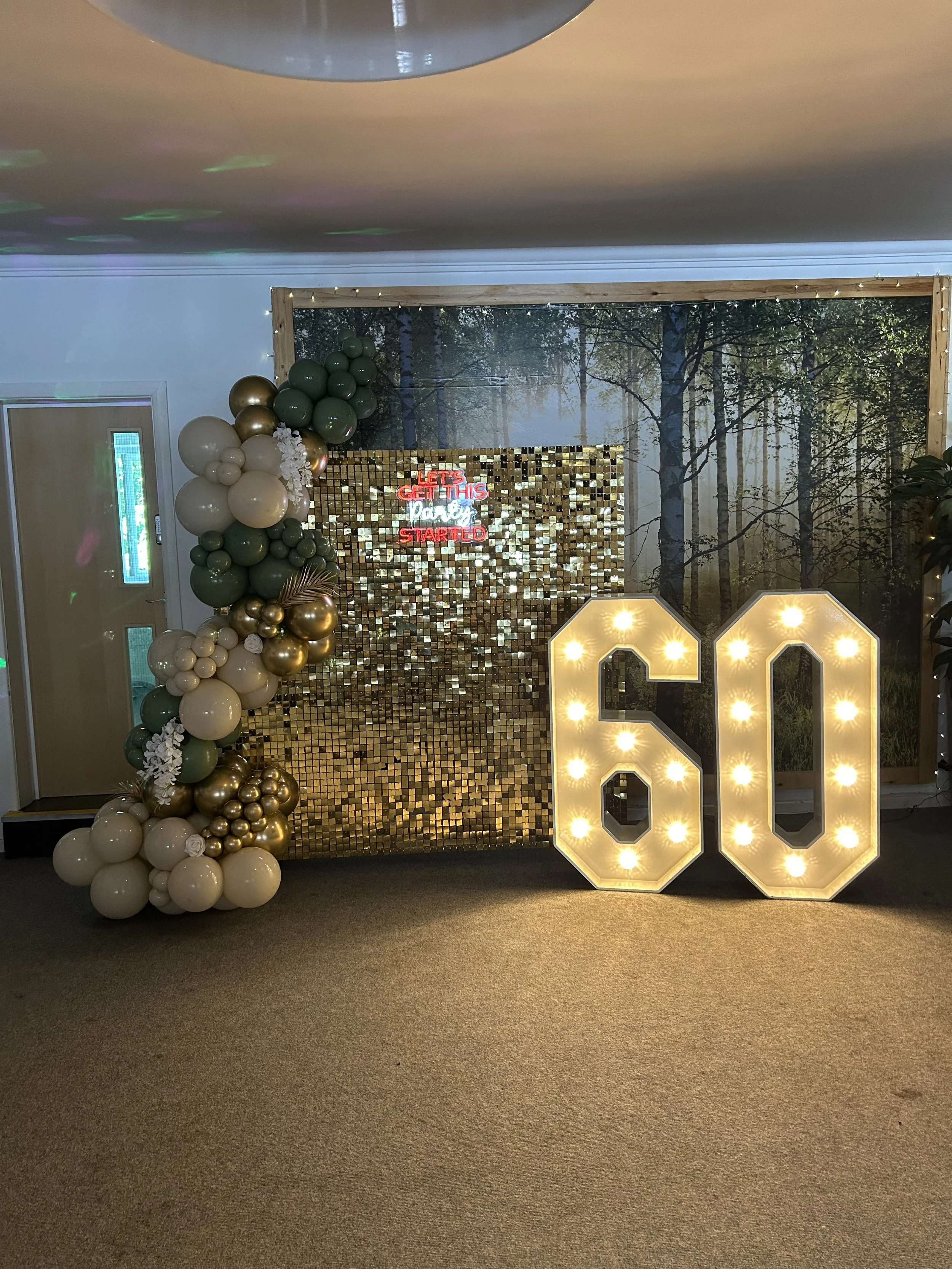 Decorative backdrop with balloons and a large lit number 60, celebrating a milestone birthday or anniversary, featuring a forest scene background, a sparkly gold mosaic wall, a neon sign, and large illuminated numbers.