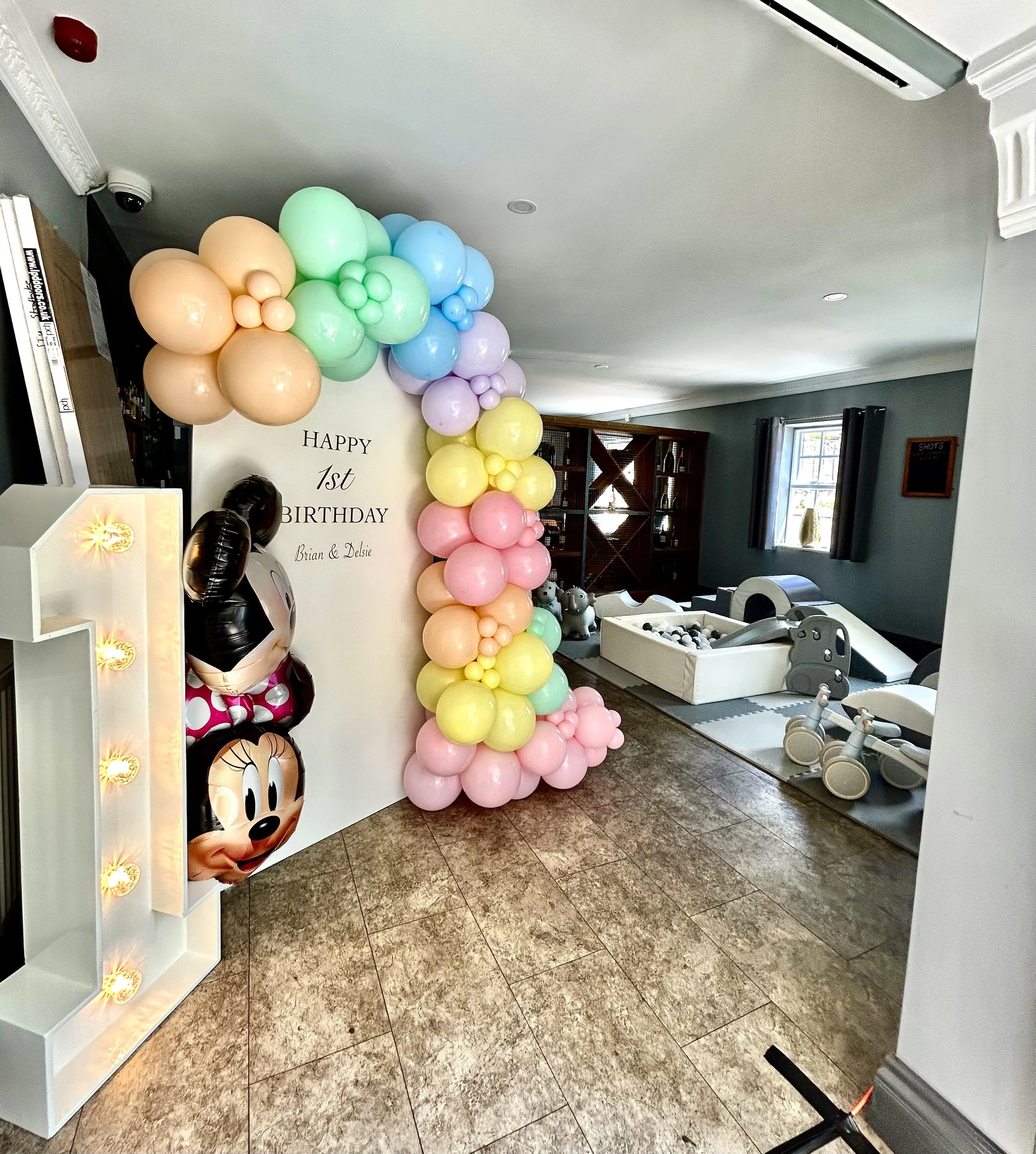 Colorful balloons arranged in an arch beside a white wall with a birthday message for a first birthday celebration. The balloons include pastel colors like yellow, pink, green, purple, and blue. Decorations include Minnie Mouse and Hello Kitty themed