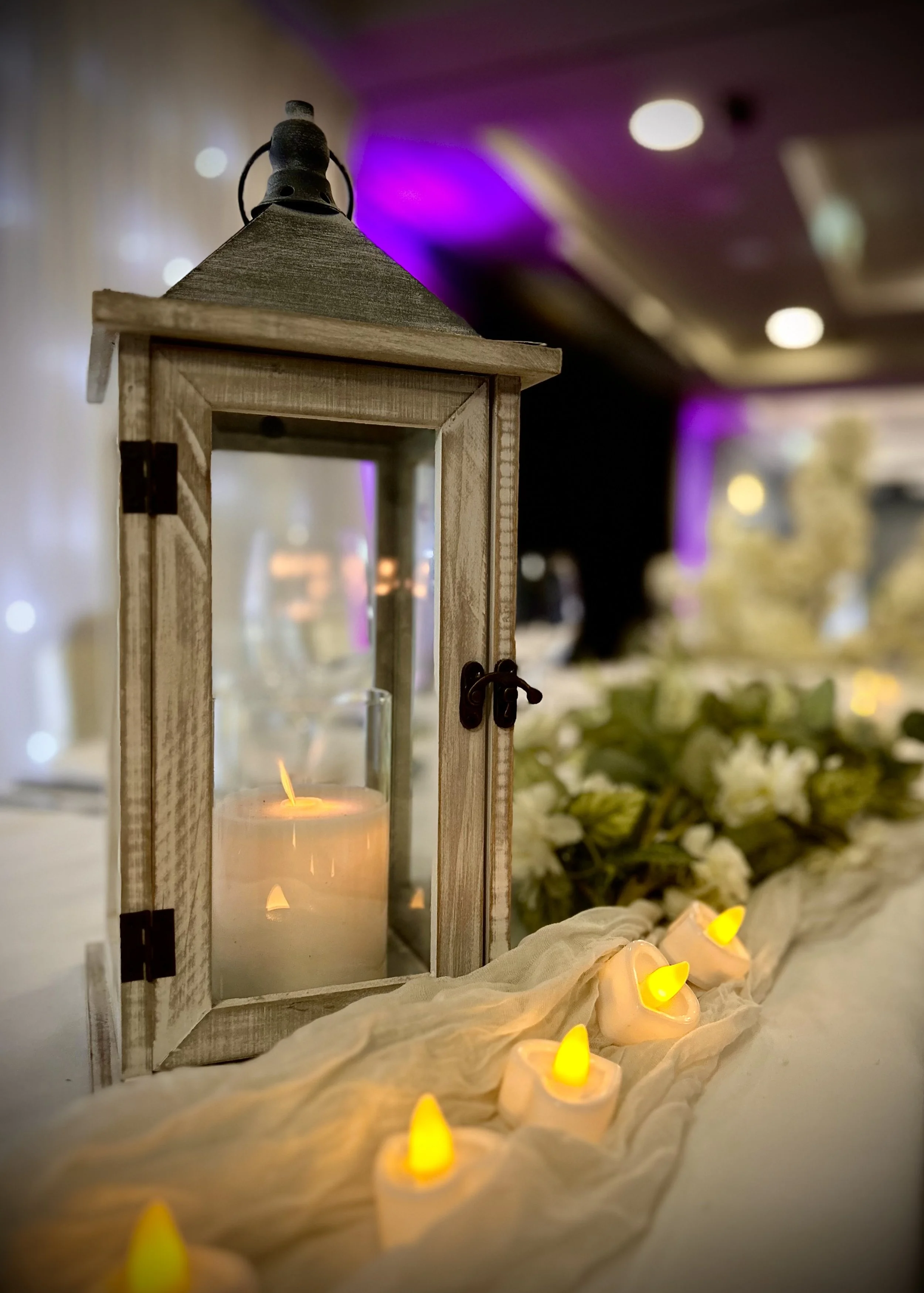 A decorative wooden lantern with a candle inside, surrounded by small yellow LED candles on white fabric, with white flowers and blurred purple and warm lighting in the background.