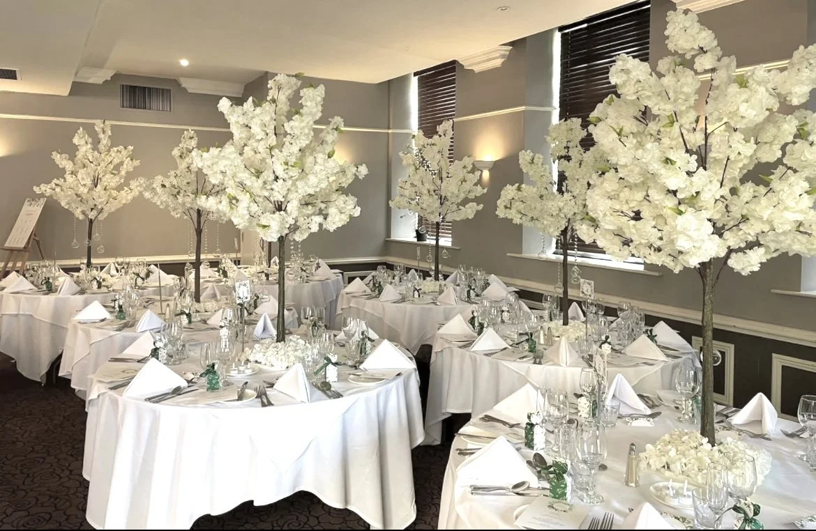 Elegant banquet hall decorated with tall white cherry blossom trees as centerpieces at each table, set for a formal event with white tablecloths, folded napkins, glassware, and silverware arranged.