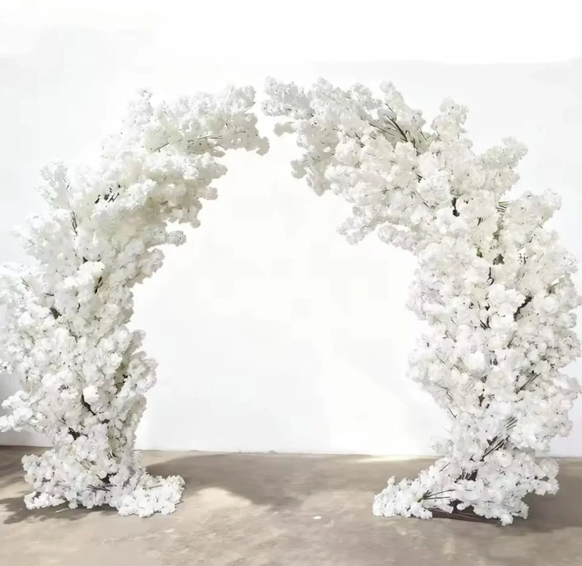 White artificial floral arch with a minimalist design, placed against a plain white wall on a gray floor.