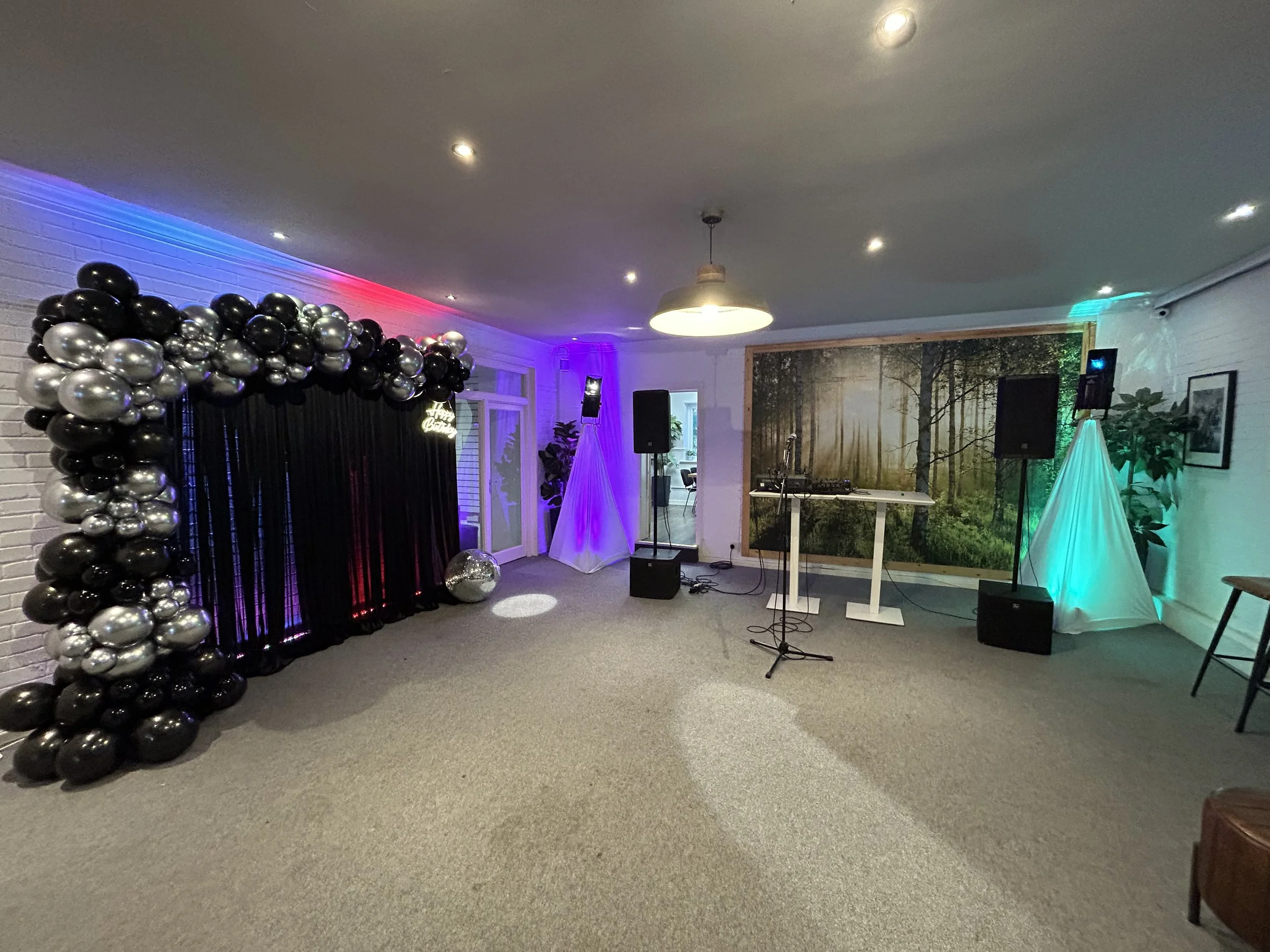 Indoor party setup with balloon arch, DJ equipment, colorful lighting, and forest-themed backdrop.