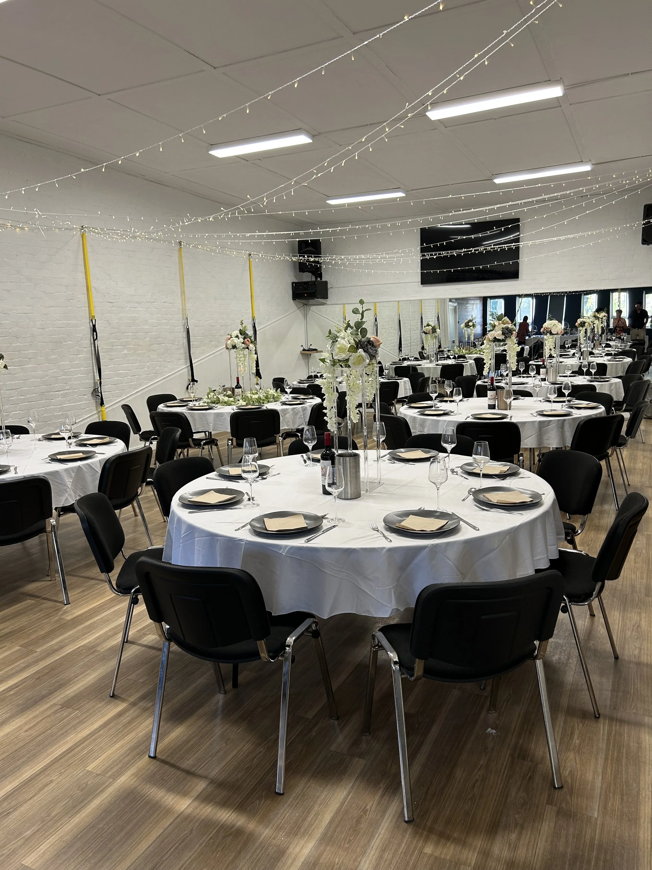 A banquet hall with round tables covered with white tablecloths, decorated with floral centerpieces, set with black plates, napkins, silverware, and wine glasses. Black chairs are arranged around the tables, and string lights are hanging from the whi