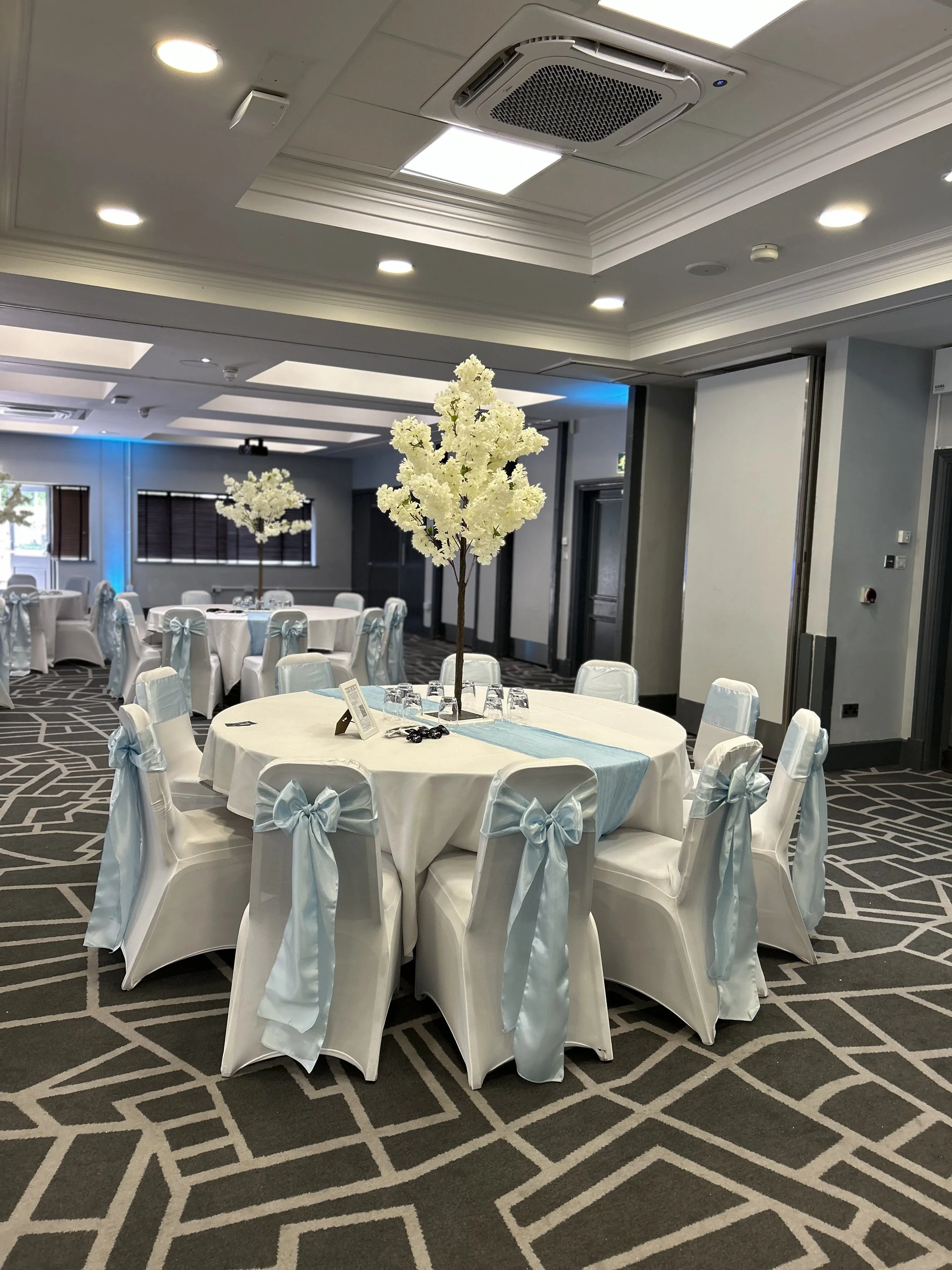 Round banquet table with white tablecloth, decorated with a light blue table runner and a tall centerpiece of white flowers. The chairs around the table are covered with white fabric and tied with light blue bows. The room has a patterned carpet, gra