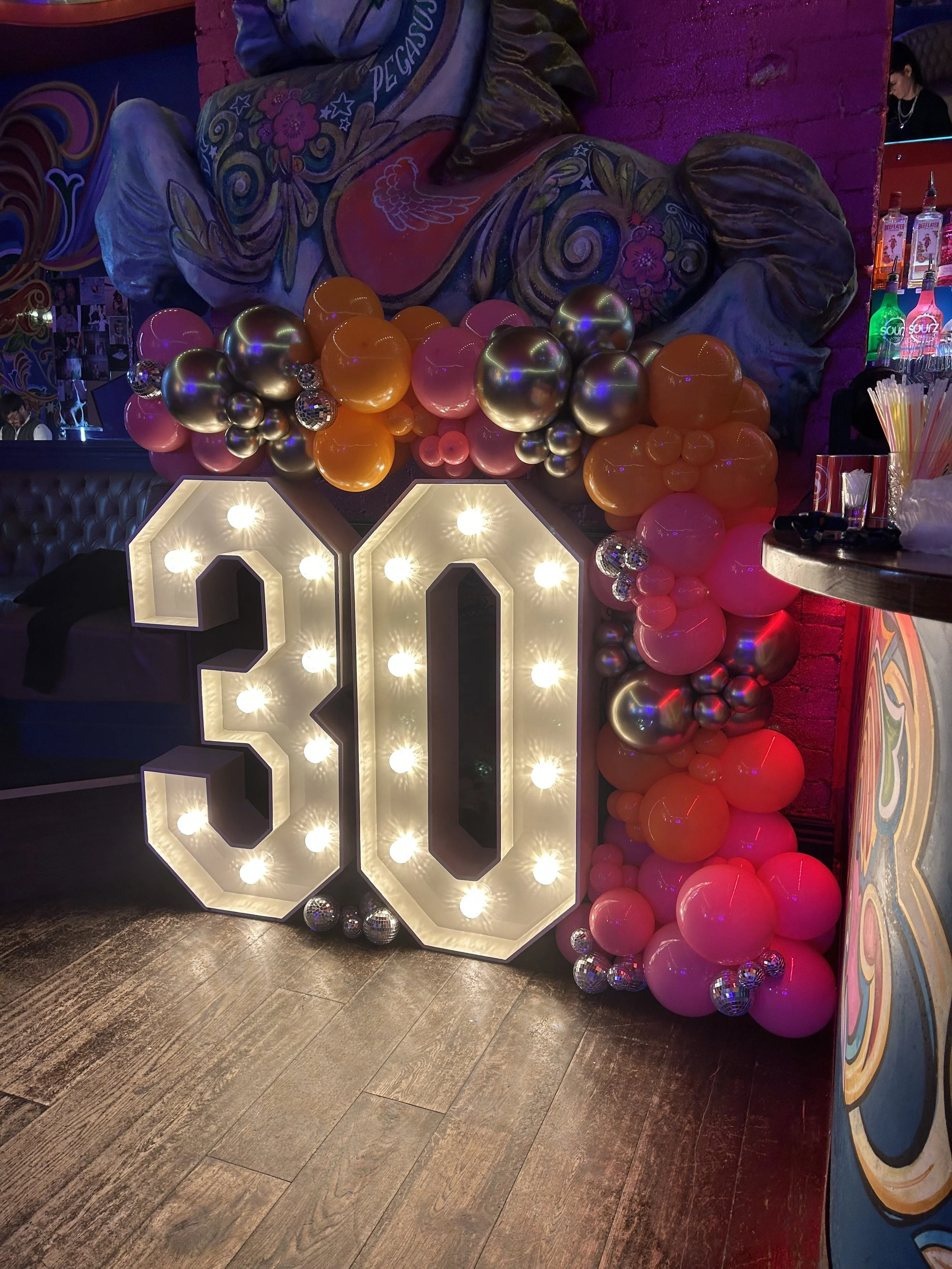 Decorative 30th birthday celebration display with large illuminated '30' sign, surrounded by colorful balloons in pink, orange, and metallic colors, against a wall with graffiti-style art and dim, colorful lighting.