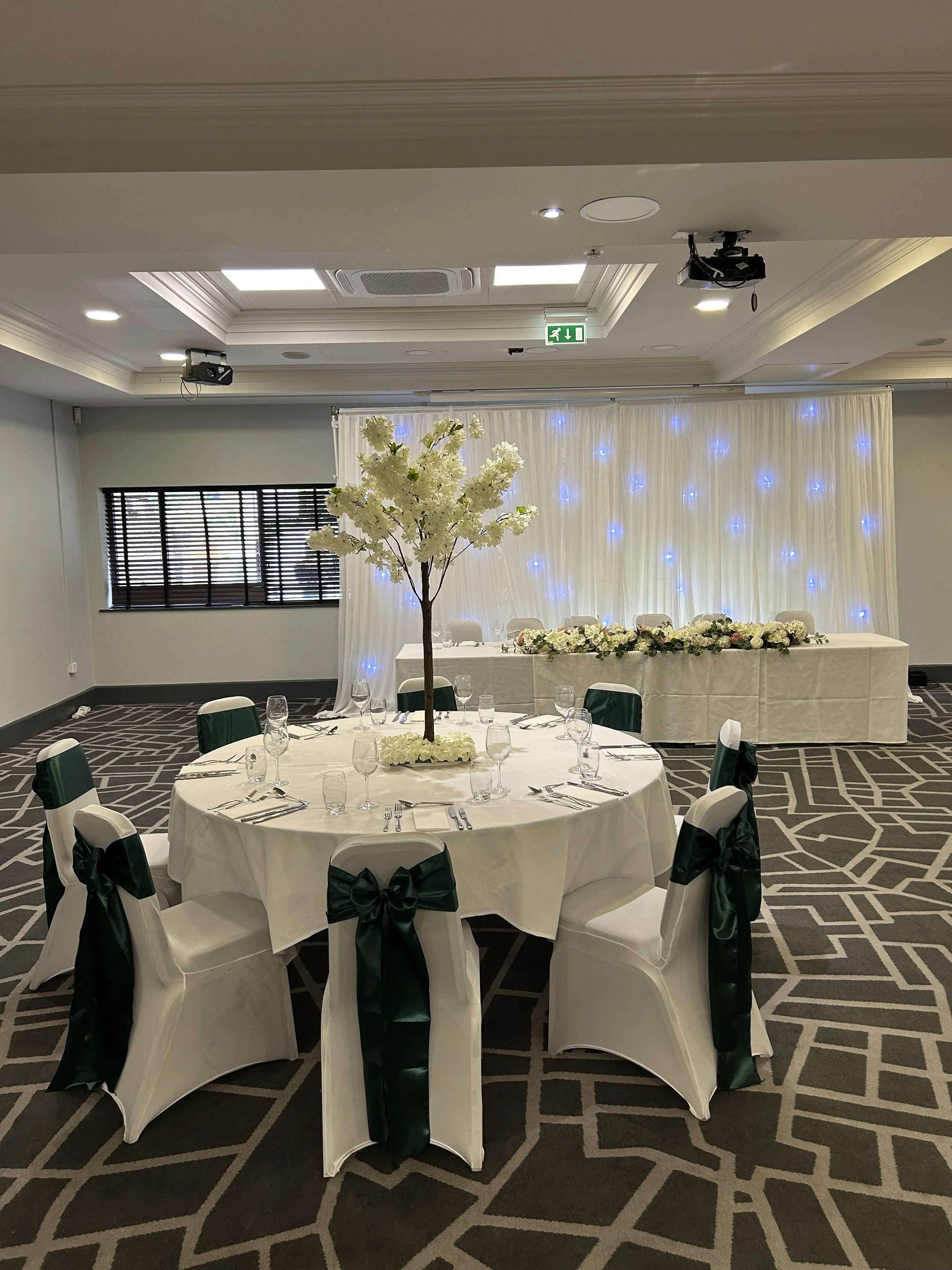 Round banquet table with white tablecloth, decorated with a tall white flower centerpiece, set with glasses and silverware. Chairs with white covers and dark green sashes surround the table. In the background, there is a long table with floral arrang
