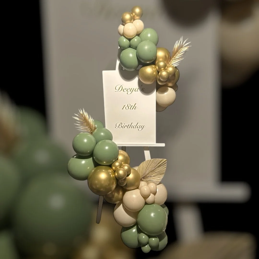 Decorative balloon arrangement featuring green, gold, and cream balloons with gold leaves, surrounding a white card that reads 'Deeya's 18th Birthday'