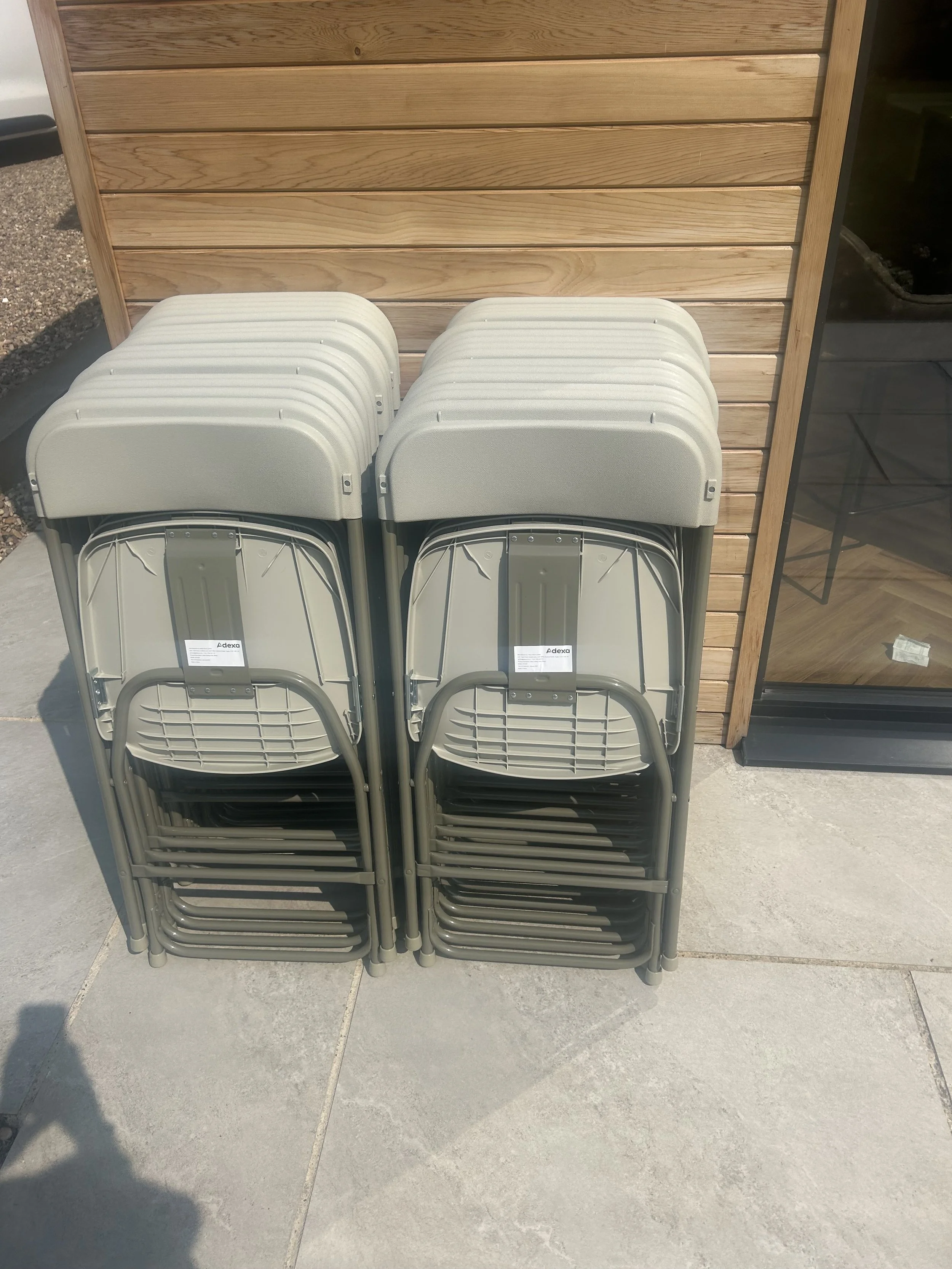 Stacked white and gray folding chairs outside on a gray tiled surface near a wooden wall.