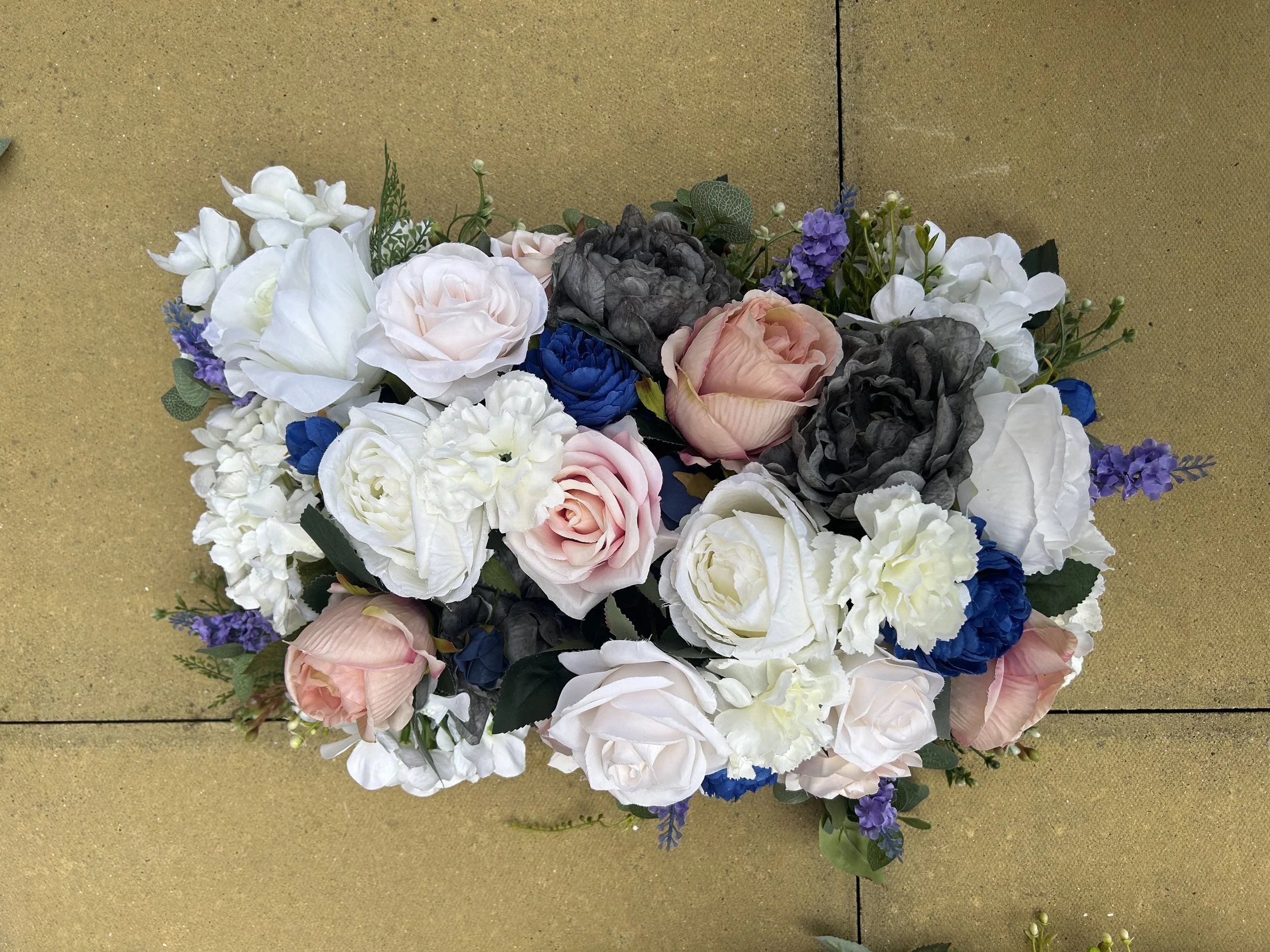 A bouquet of various artificial flowers including roses, peonies, and smaller blossoms in colors such as white, pink, blue, gray, and purple, arranged on a beige tiled surface.