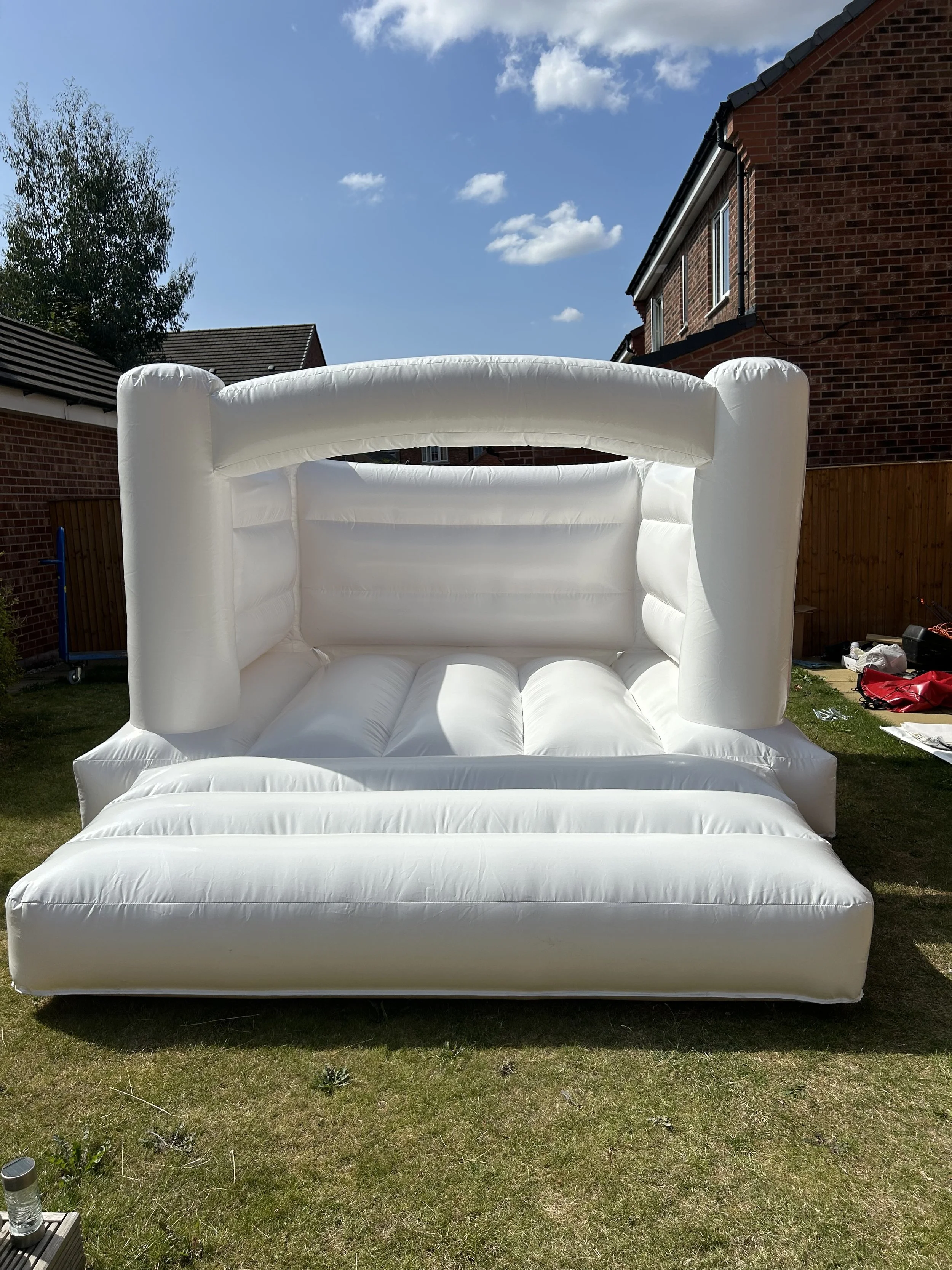 Inflatable white sofa with a high backrest and armrests, set up on grass in a backyard with brick houses and a blue sky with clouds in the background.