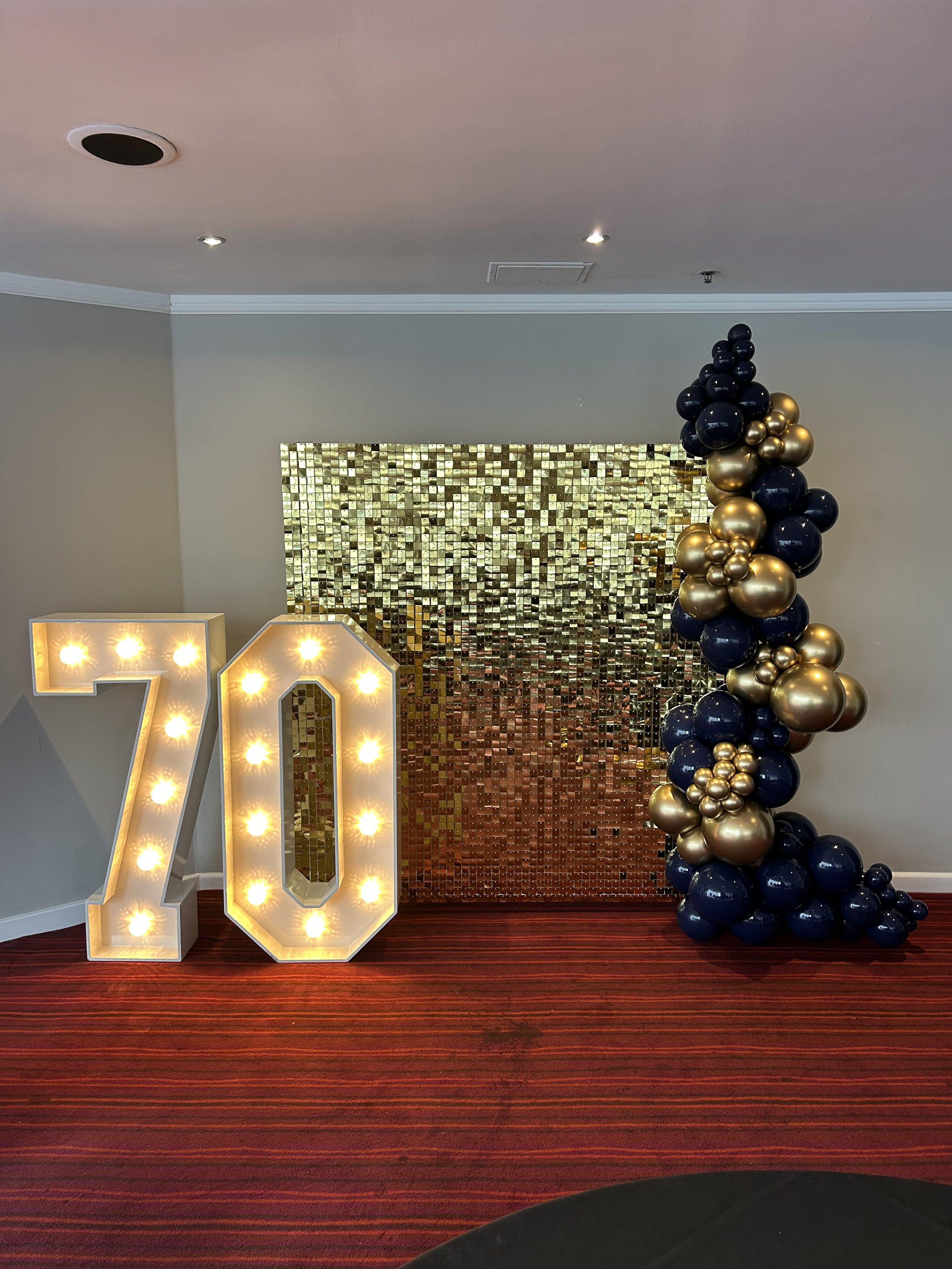 Decorative display with large illuminated number '70', a gold and navy balloon arrangement, and a shiny gold mosaic backdrop on a red carpeted floor.