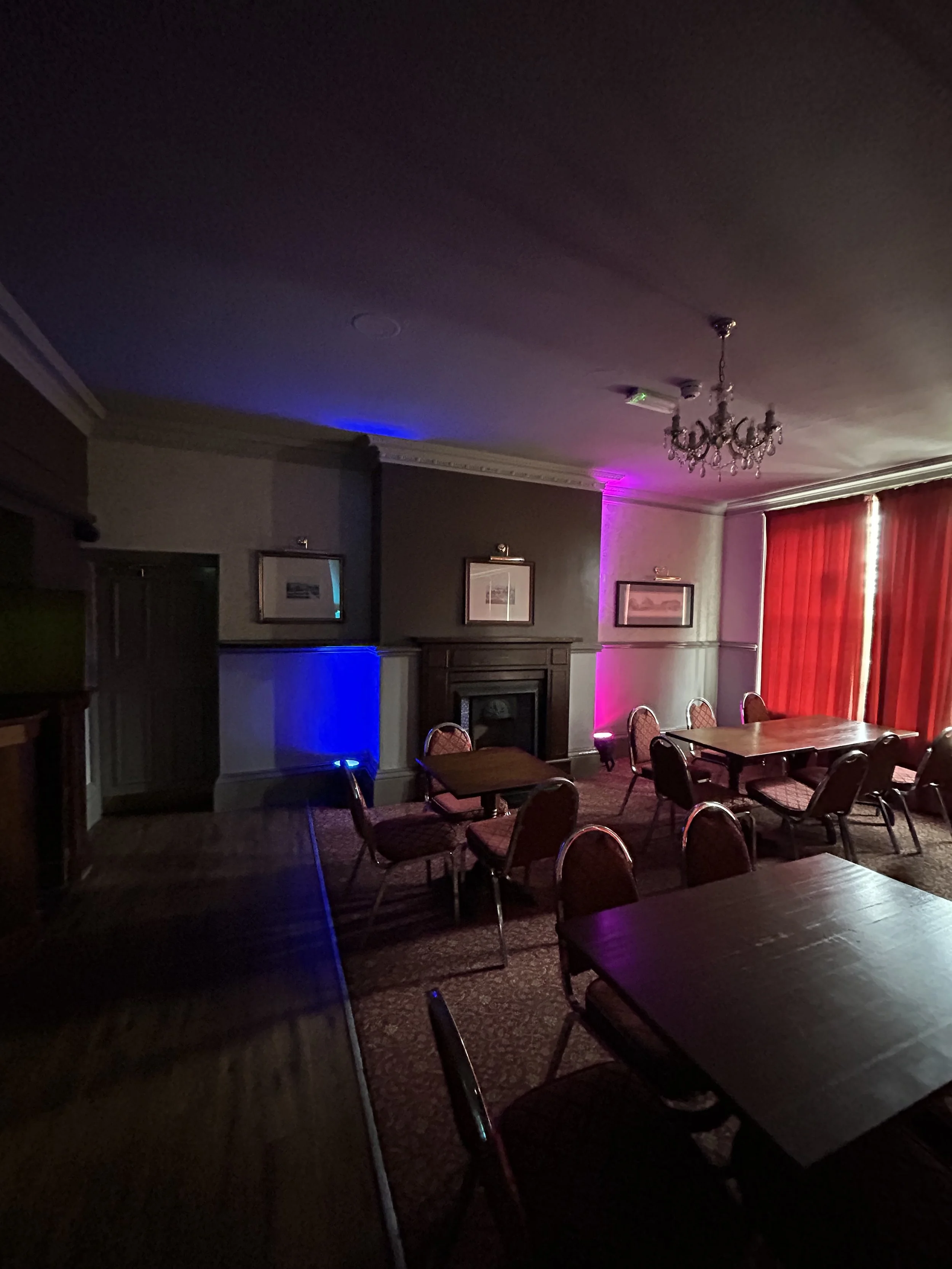 A dimly lit banquet room with tables and chairs, red curtains, a chandelier, and colored lights illuminating the walls.