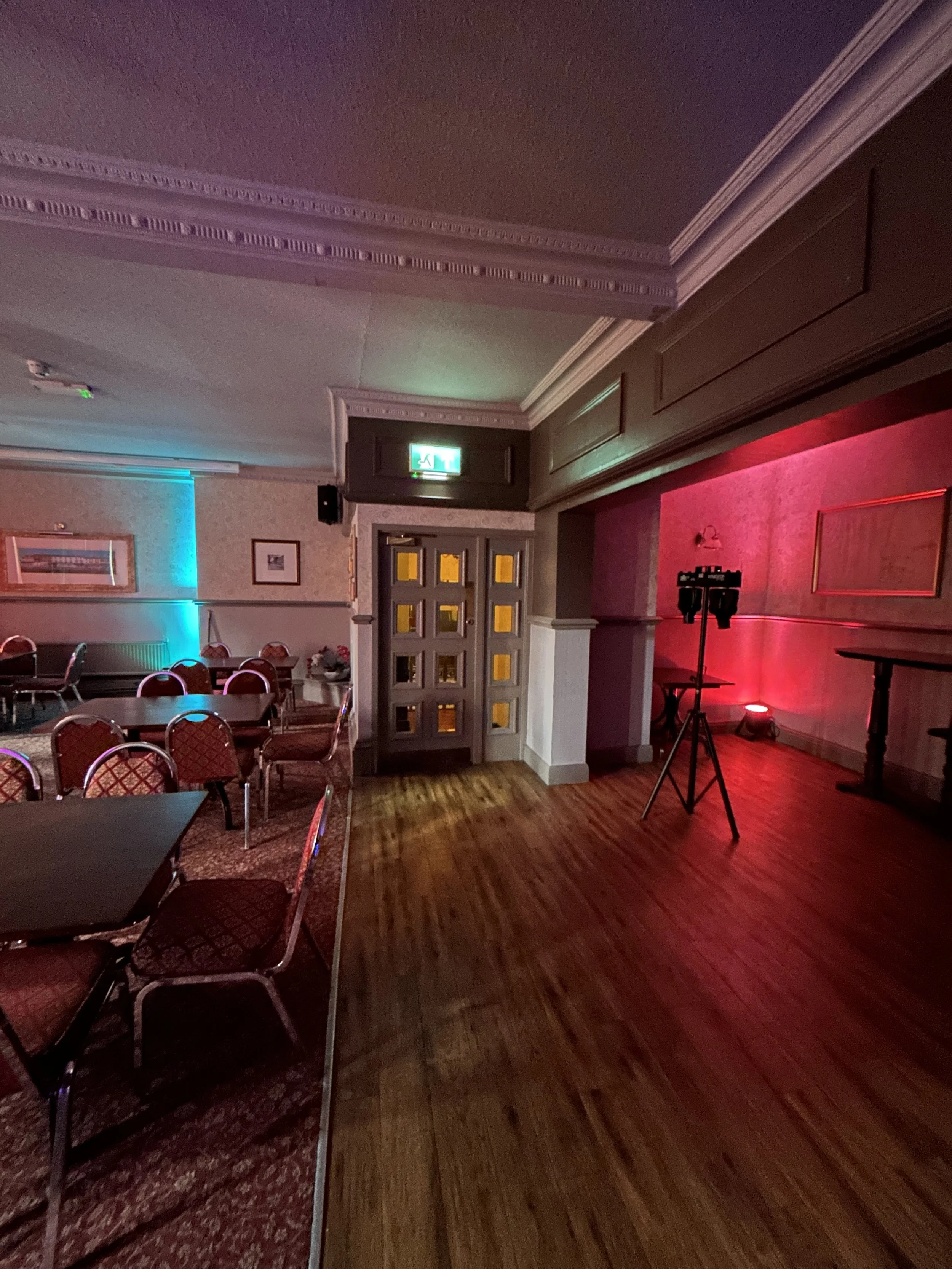 An empty event room with tables and chairs on the left, colorful LED lighting, a wooden floor, and a dance floor area on the right with lighting equipment.