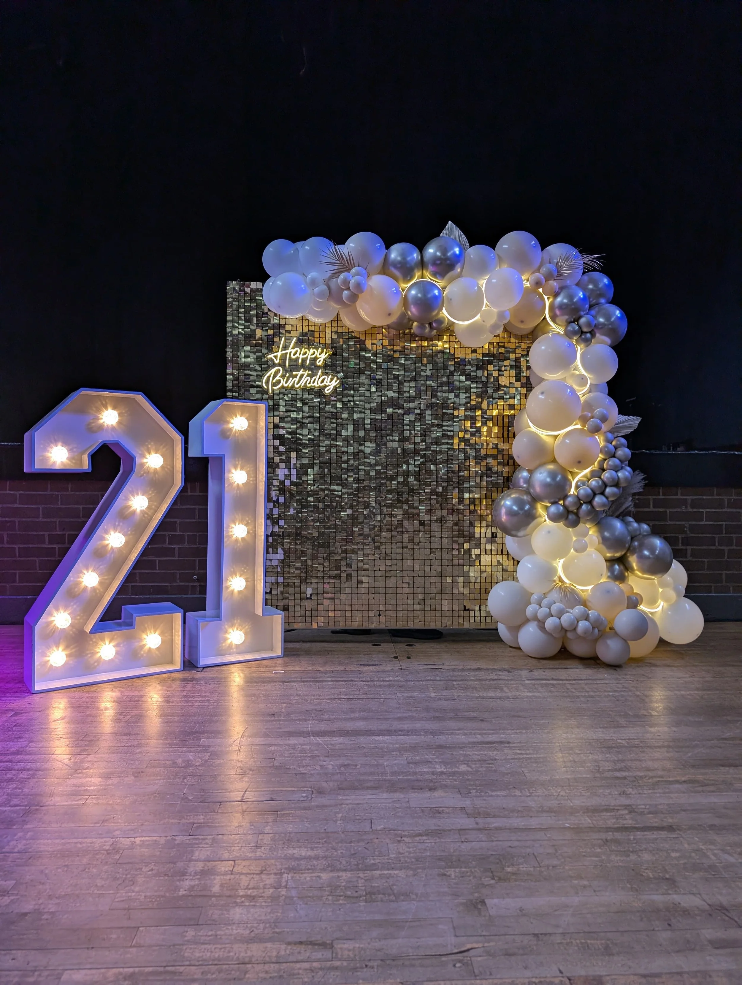 Decorative birthday backdrop with a large illuminated number 21, a cluster of white and metallic balloons, and a small neon sign that reads 'Happy Birthday', set against a dark wall with a reflective gold mosaic panel.