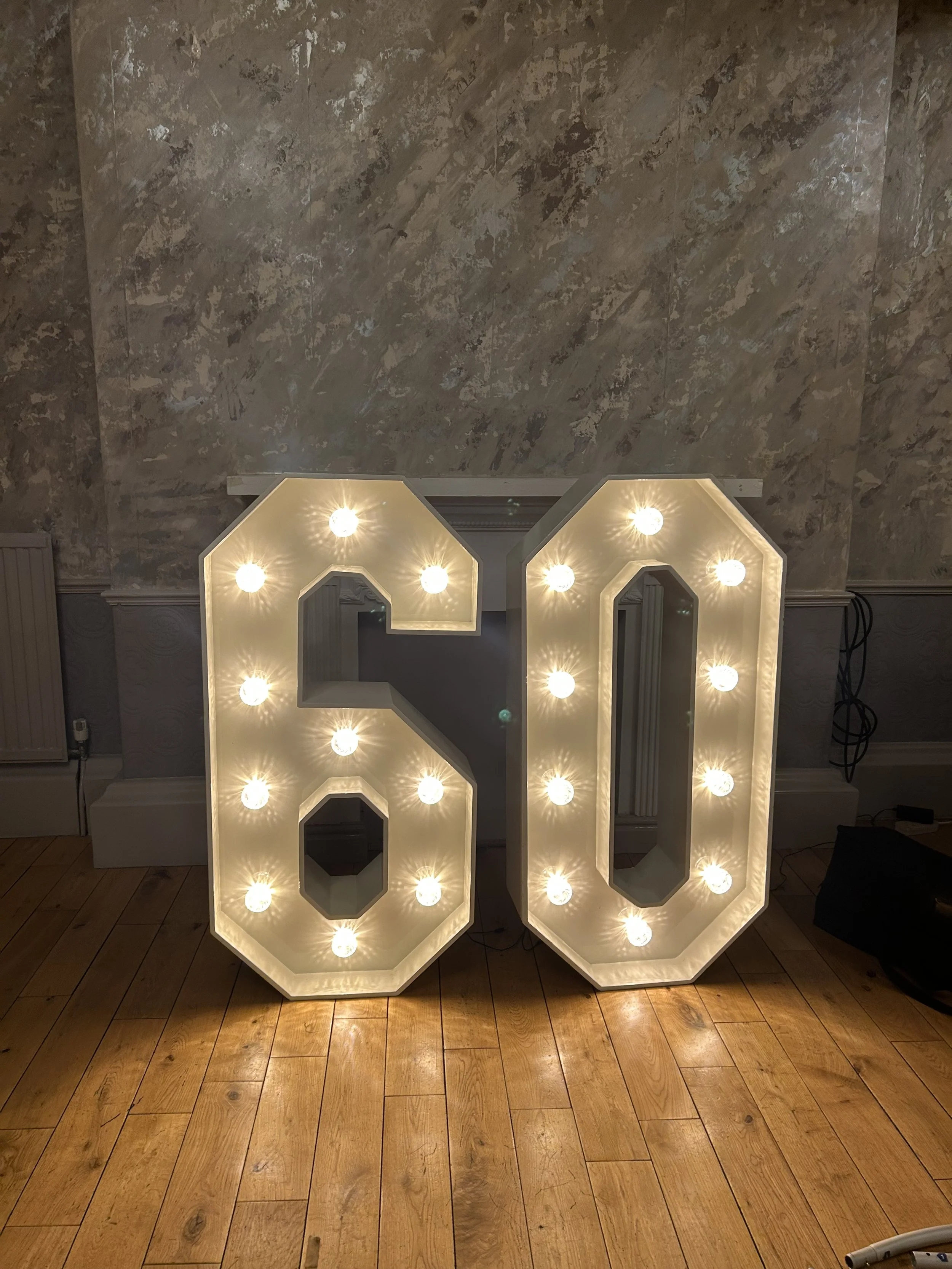 Large illuminated marquee sign spelling out the number 60 with white light bulbs, placed on a wooden floor against a textured wall.