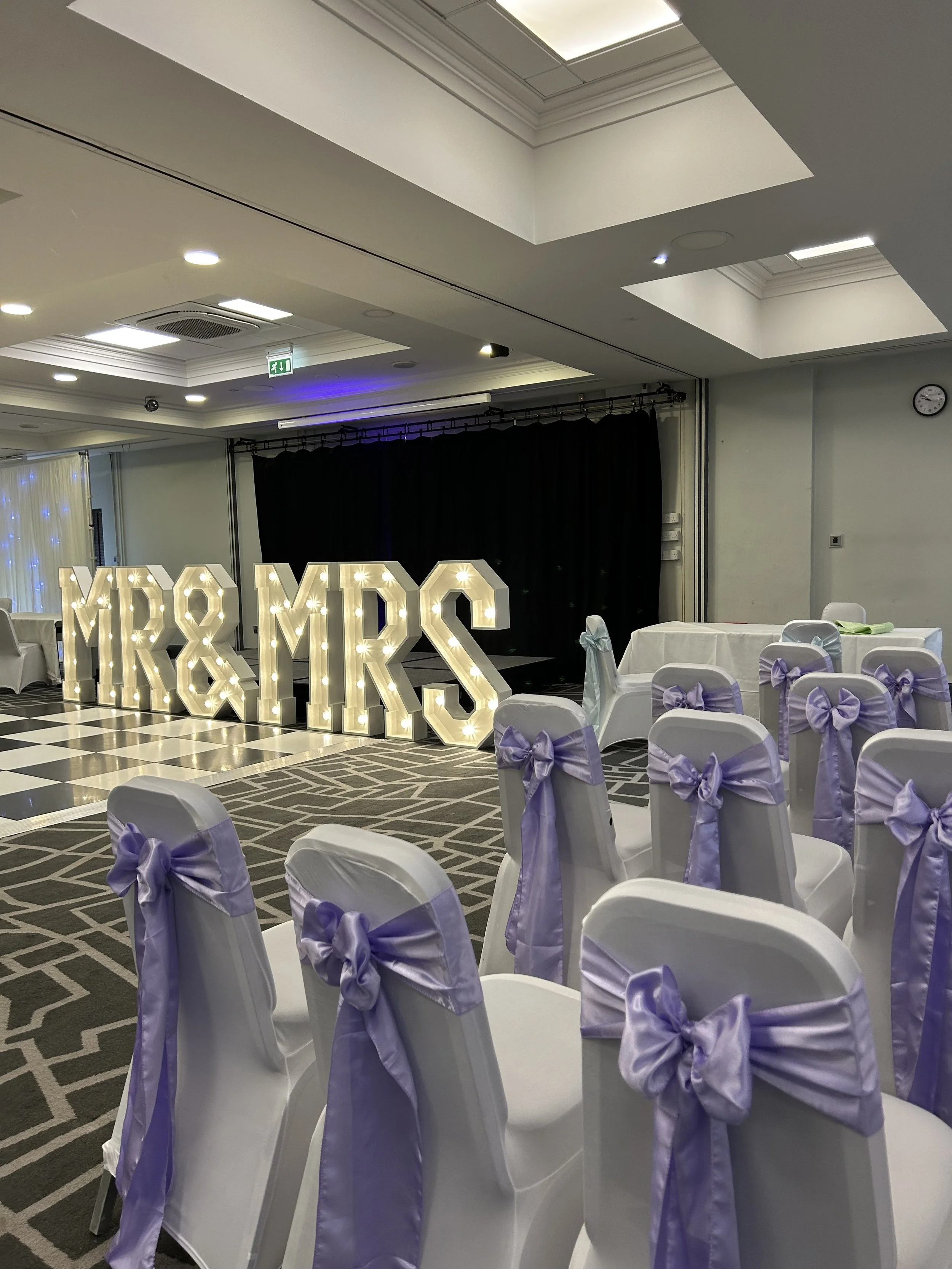 Event space decorated with illuminated letters spelling 'MR & MRS', surrounded by chairs with white covers and lavender bows, and black curtains in the background.