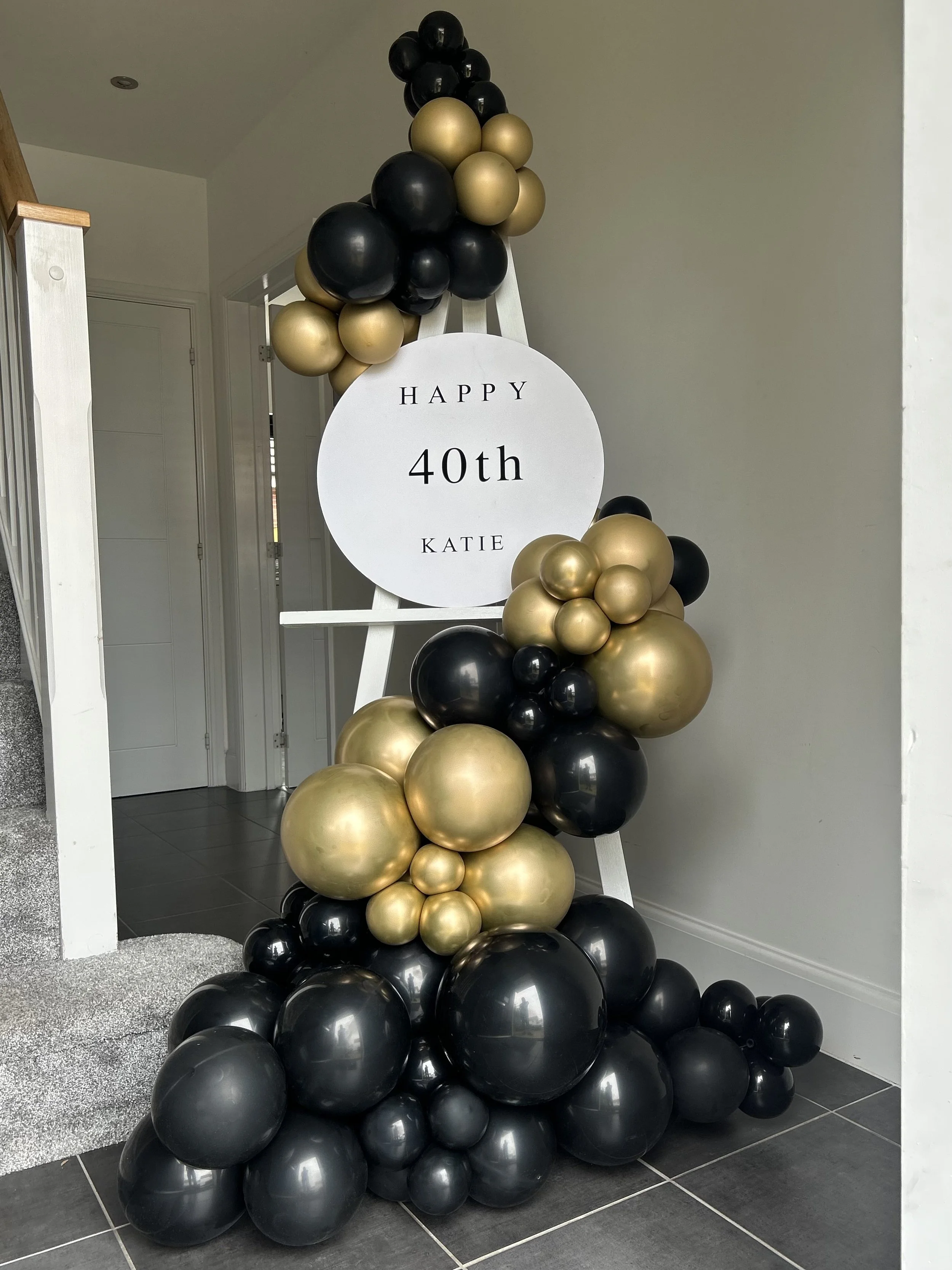 Balloon decoration with black and gold balloons surrounding a white circular sign that reads 'Happy 40th Katie' on an easel.
