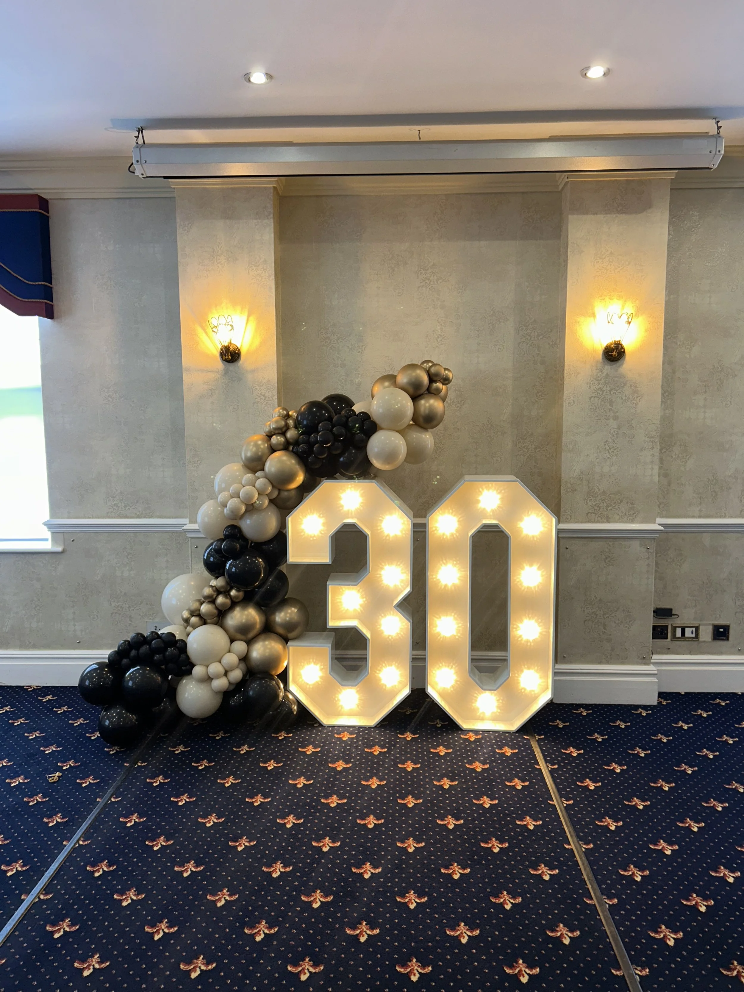 Large illuminated number 30 surrounded by black, white, and gold balloons, decorated for a celebration.