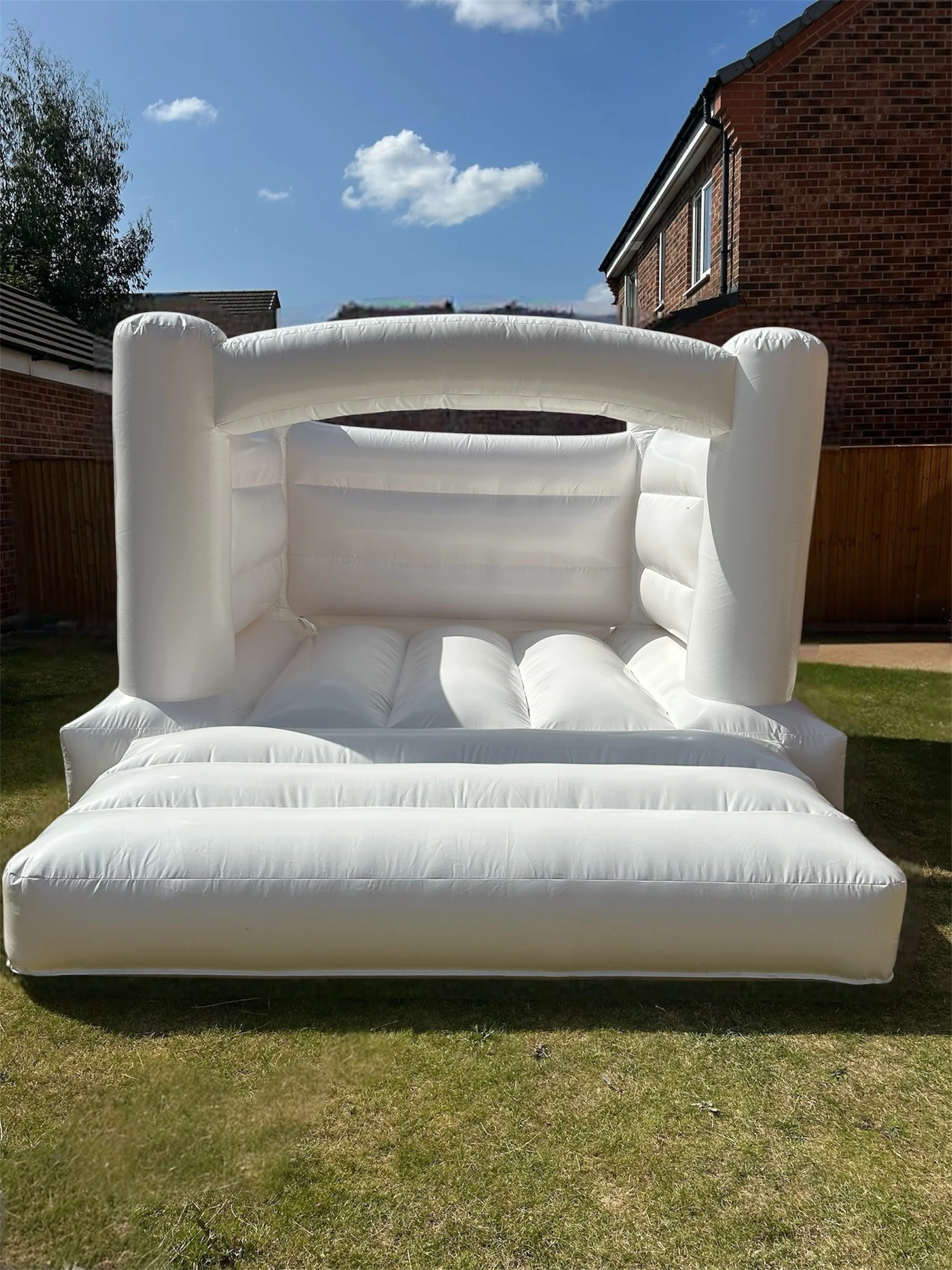 Inflatable white outdoor lounge chair on a grass lawn in a residential backyard with brick houses, a wooden fence, and a partly cloudy blue sky.