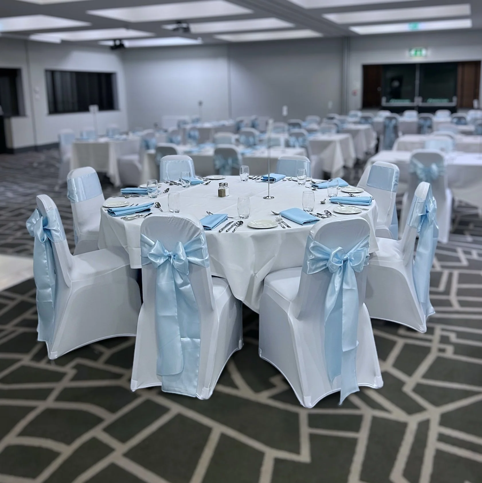 Round banquet table set for a formal event with white tablecloths, blue napkins, glassware, plates, silverware, and white chairs decorated with blue bows, in a large event hall.
